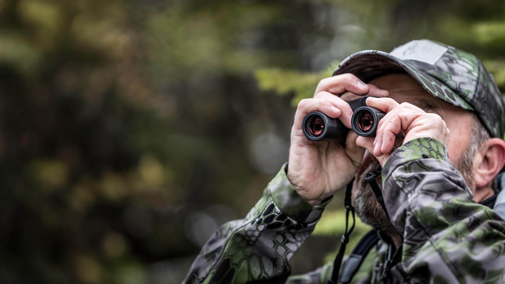 A man in camouflage clothing is observing through ZEISS binoculars in a forested area.