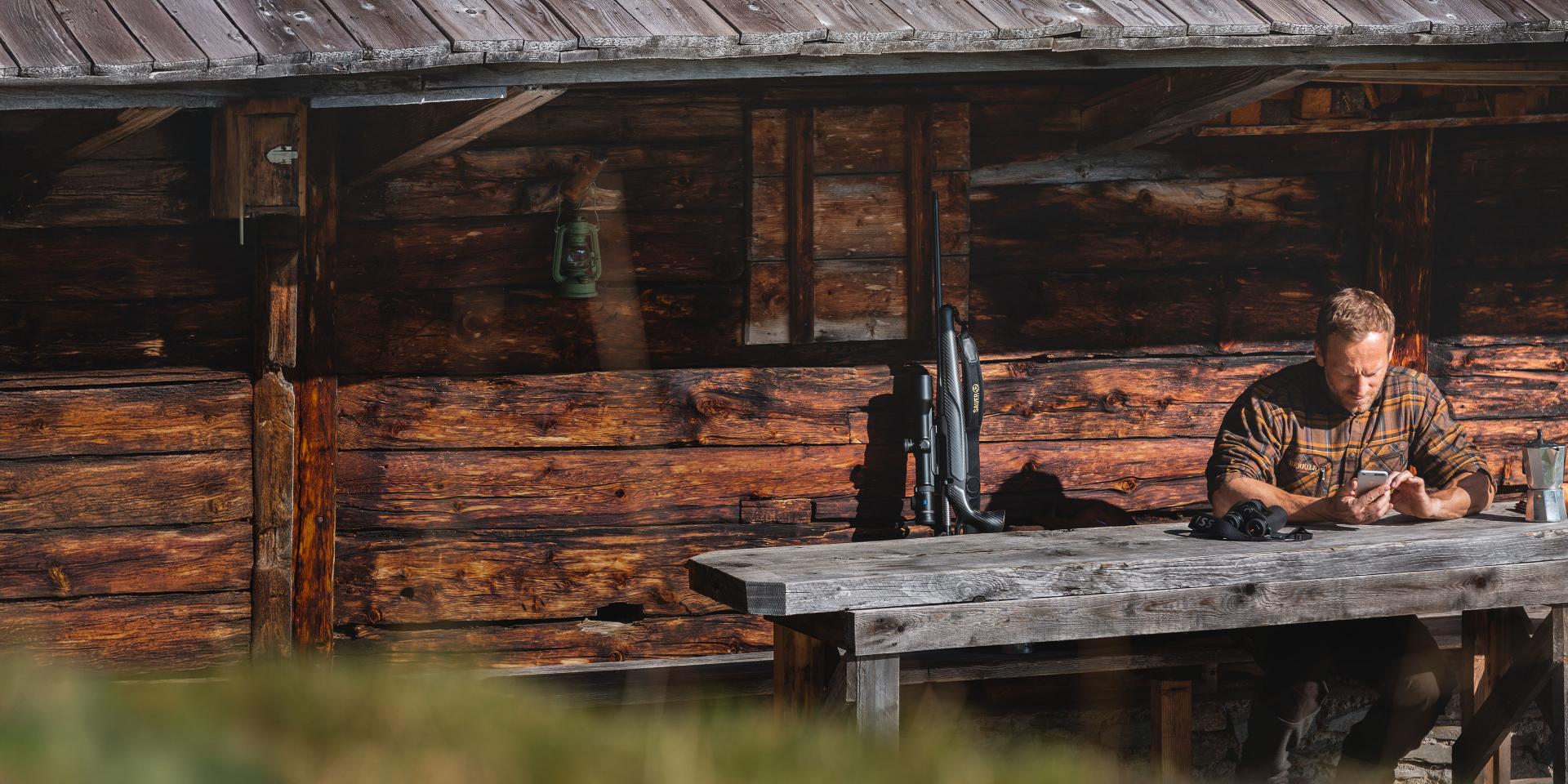 A man sits at a rustic wooden table outside a log cabin, examining ZEISS optics equipment in natural sunlight.