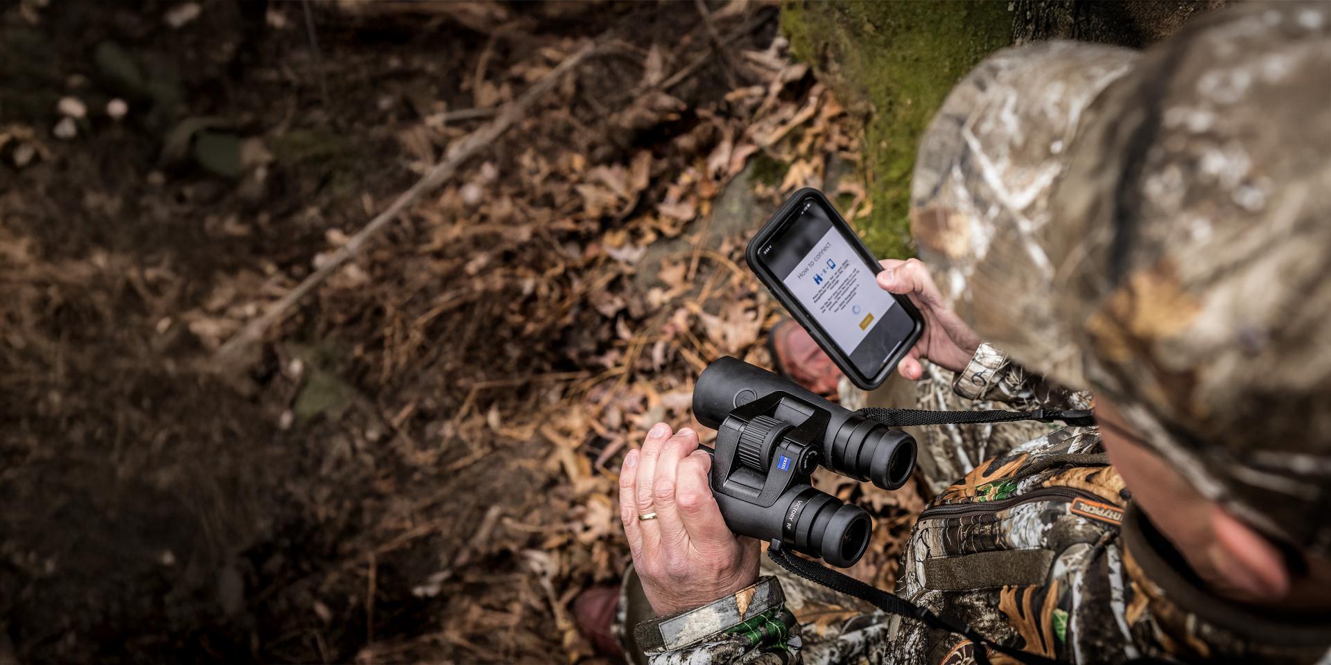 A person in camouflage gear holds a ZEISS binocular and a smartphone while standing in a forested area with dry leaves on the ground.