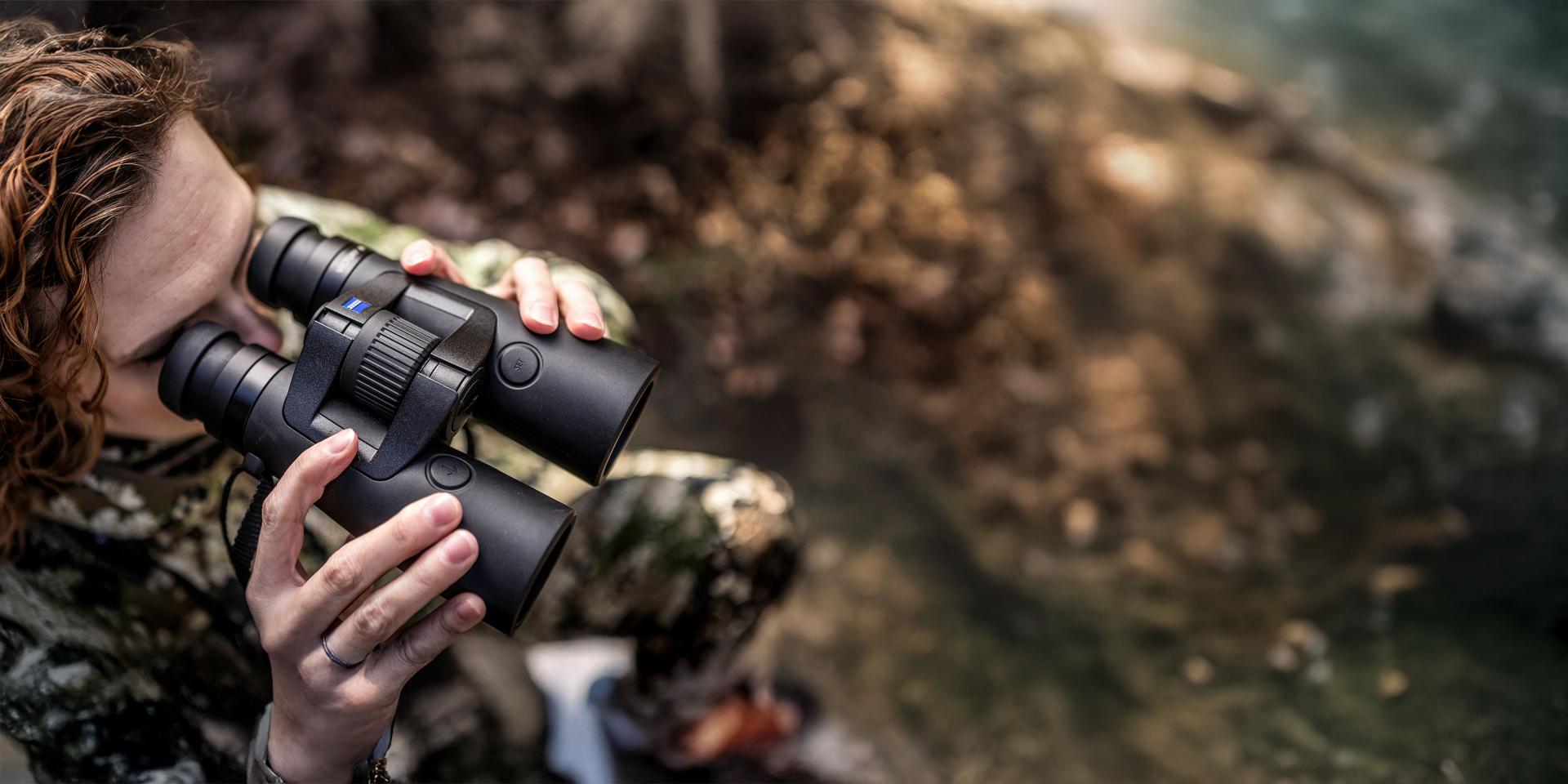 Person in camouflage clothing using ZEISS binoculars while observing nature near a body of water.