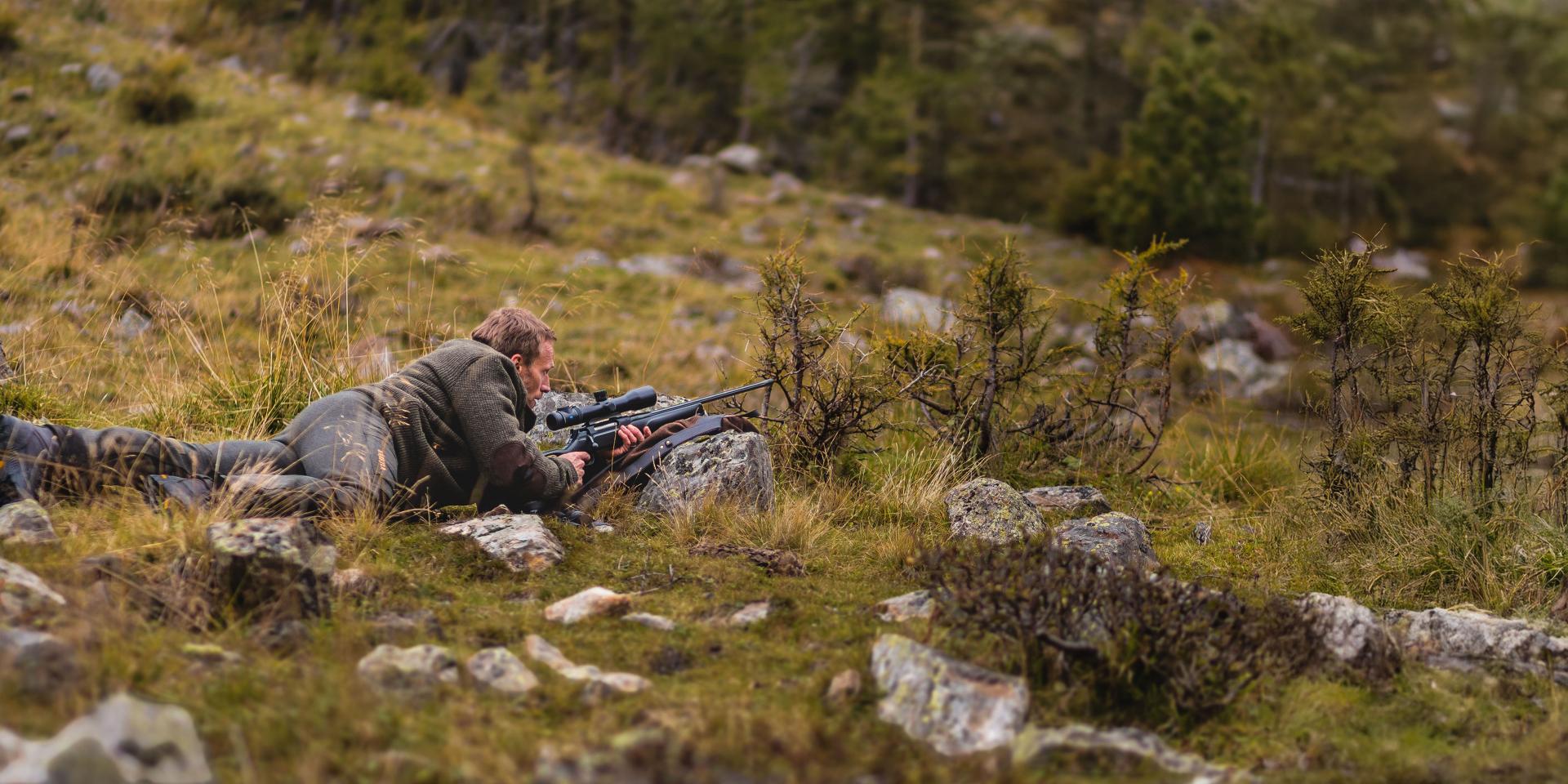 A person lies prone in a rocky, grassy landscape aiming a rifle equipped with a ZEISS scope.