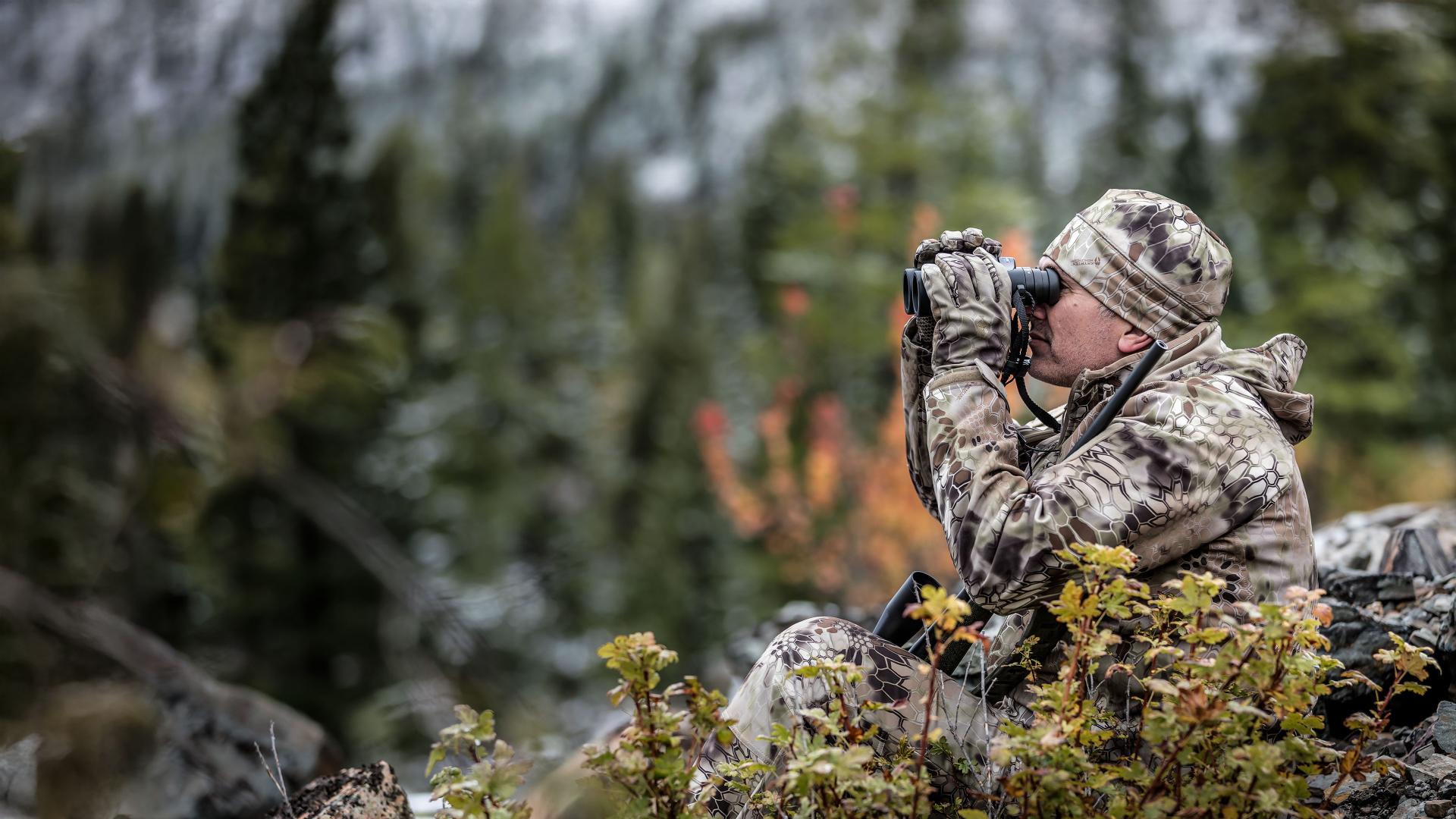 A person in full camouflage gear is using ZEISS binoculars while seated in a rugged, forested mountain landscape.