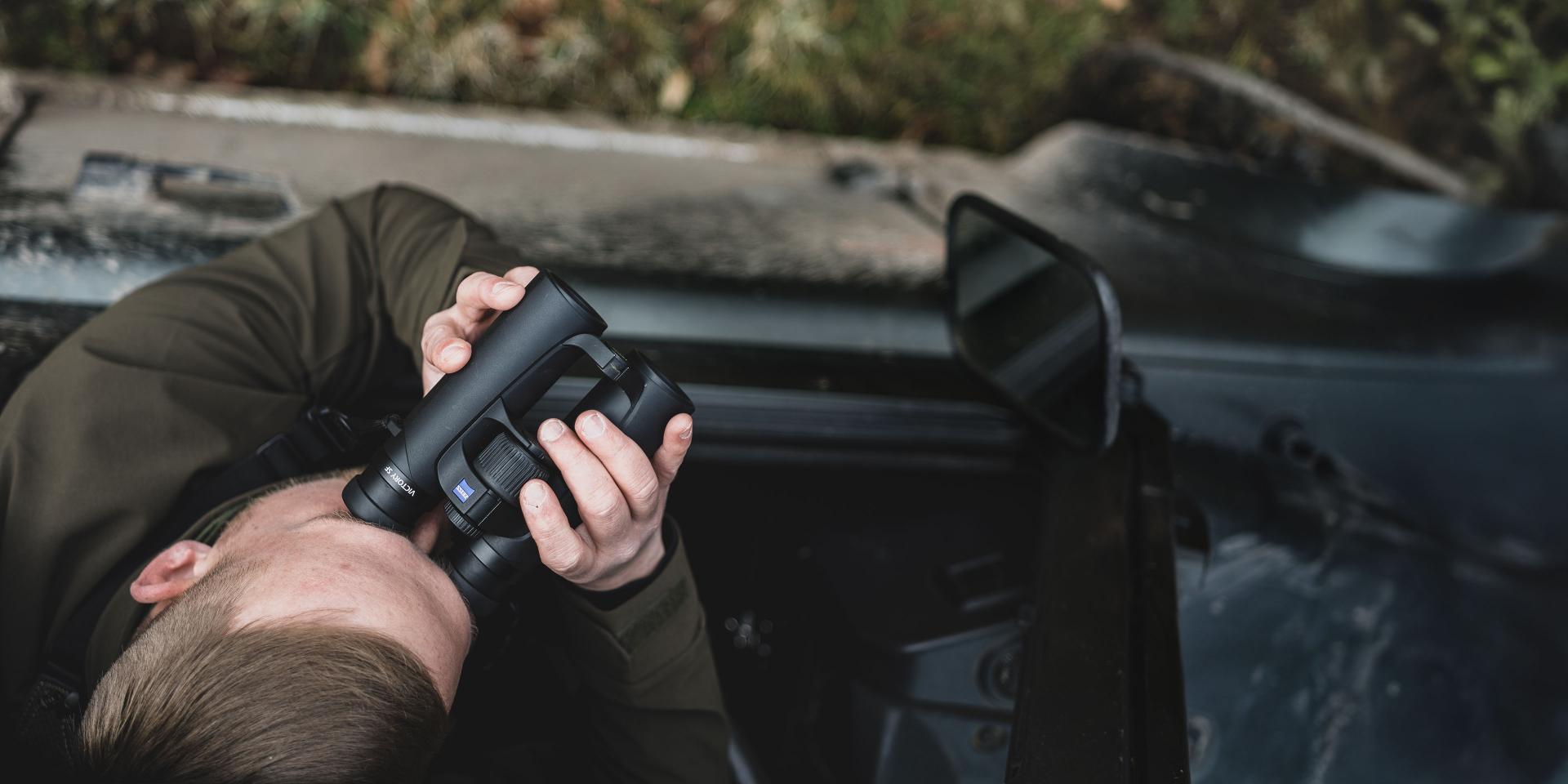 Person looking through a pair of ZEISS binoculars while leaning out of a vehicle window.