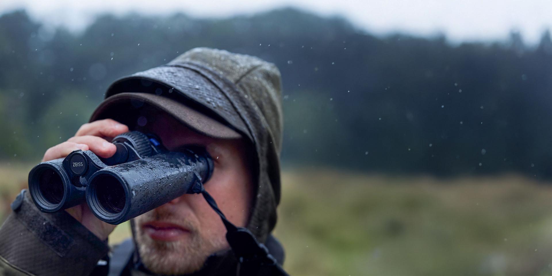 A person wearing a hooded jacket uses ZEISS binoculars in a rainy outdoor setting.