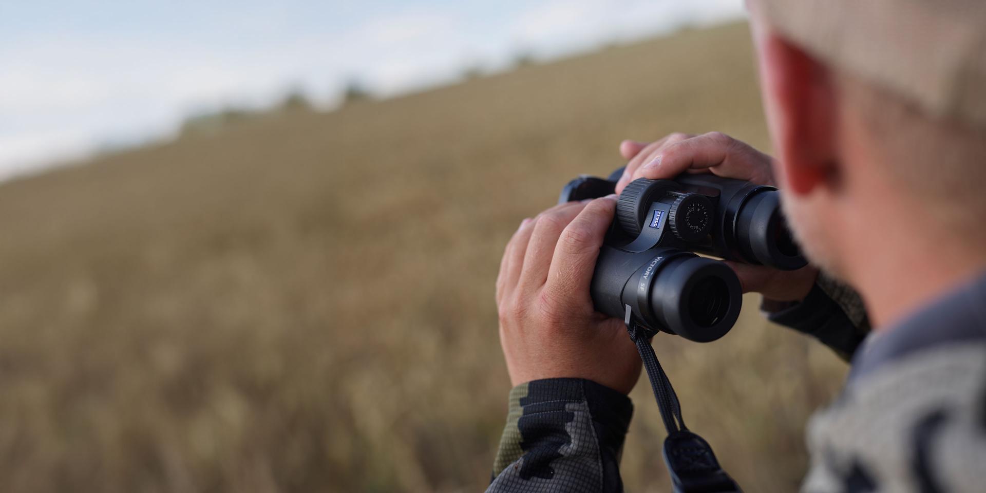 Person using ZEISS binoculars to observe a wide open field during daytime.
