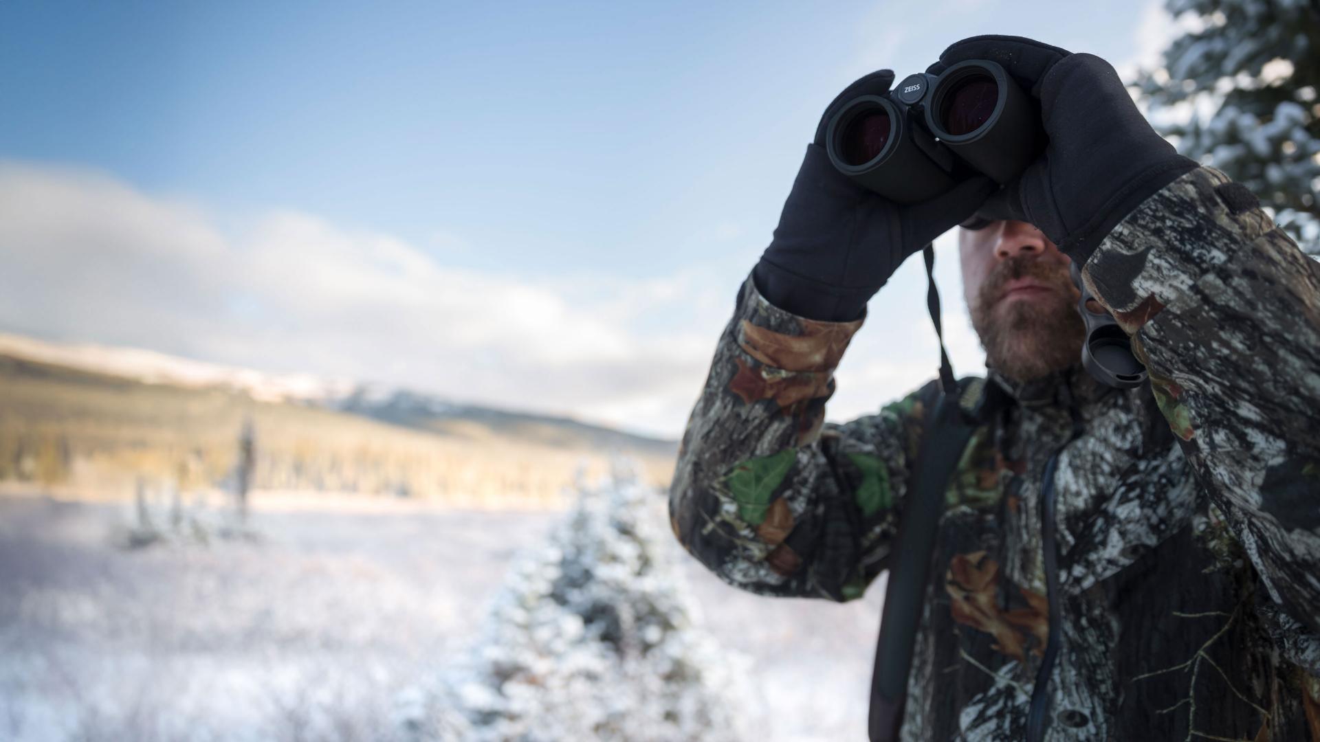 Man in camouflage clothing using ZEISS binoculars to observe a snowy landscape with mountains and trees in the background.