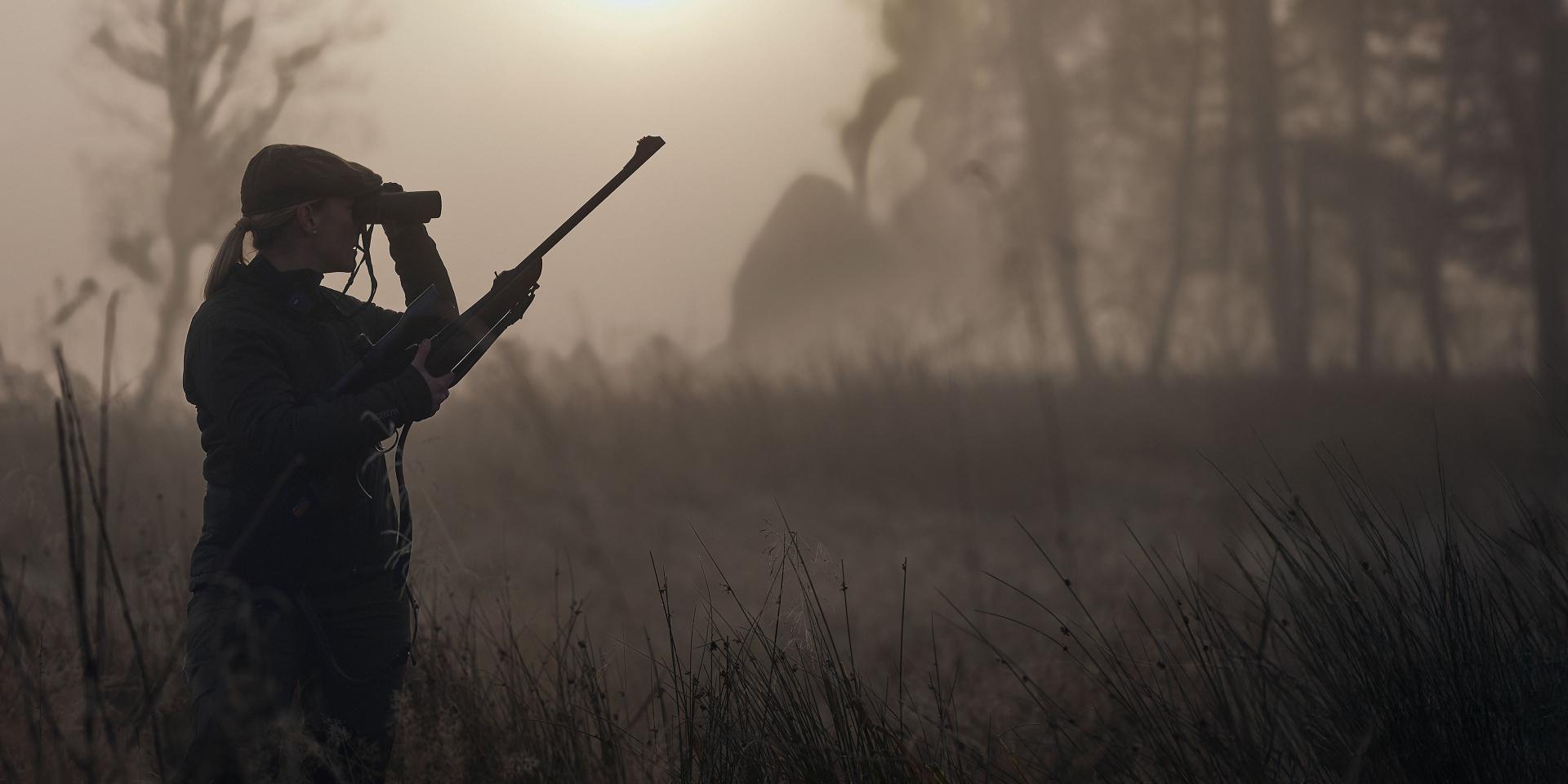 Hunter using ZEISS binoculars and holding a rifle in a foggy, early morning forest landscape.