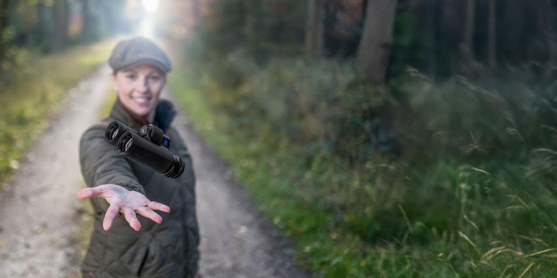 A person outdoors on a forest path is tossing a pair of ZEISS binoculars towards the camera.