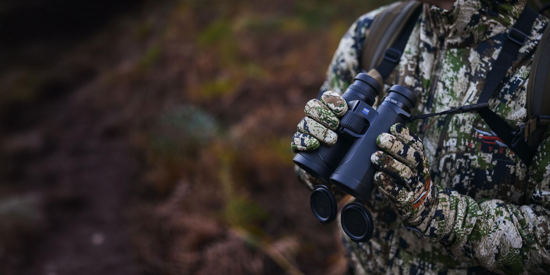 Person in camouflage gear holding a pair of ZEISS binoculars in an outdoor setting.