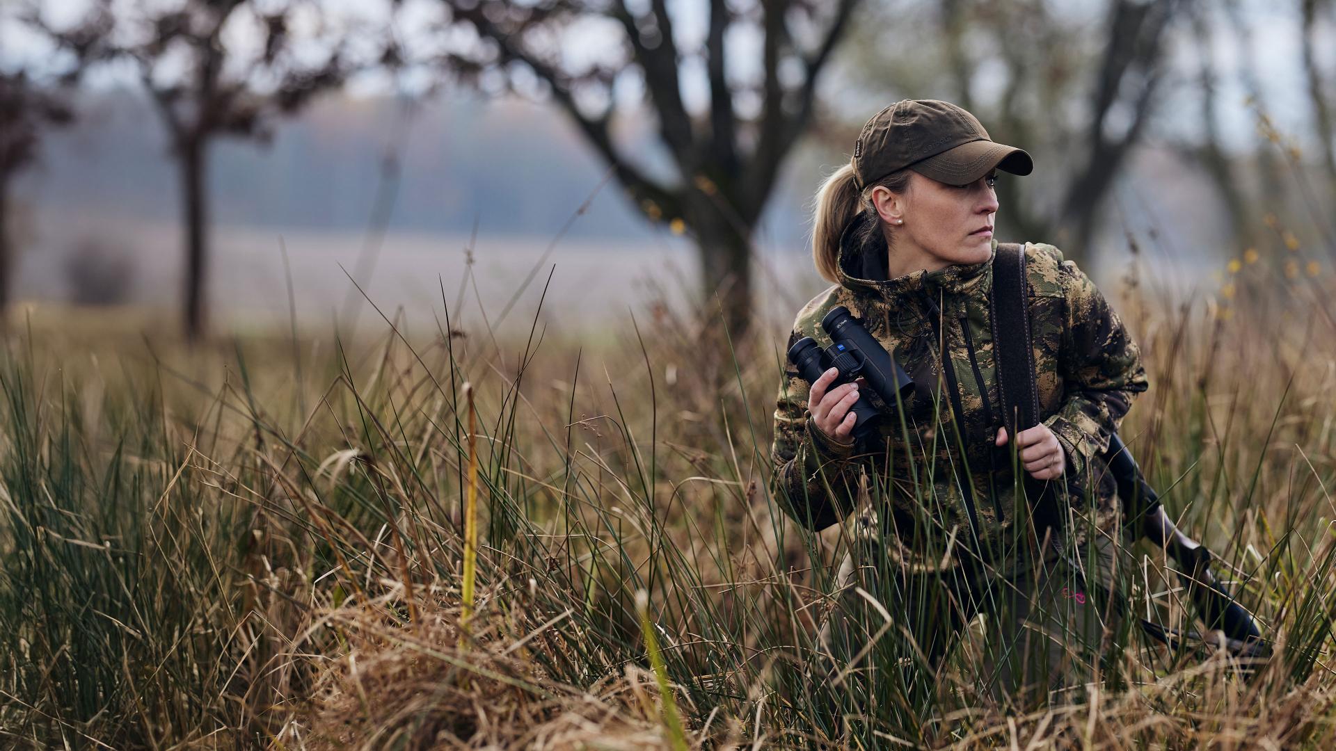 A woman in camouflage gear stands in tall grass holding a ZEISS binoculars and a rifle in a woodland setting.