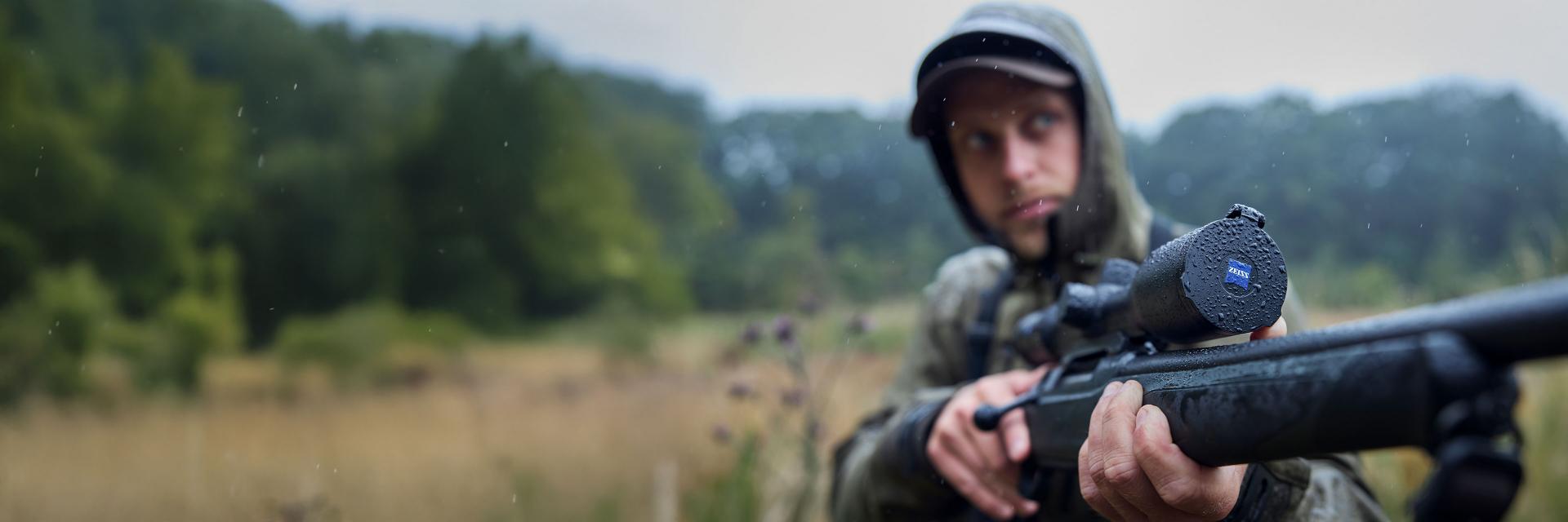 A person holding a rifle equipped with a ZEISS riflescope in a forest setting.
