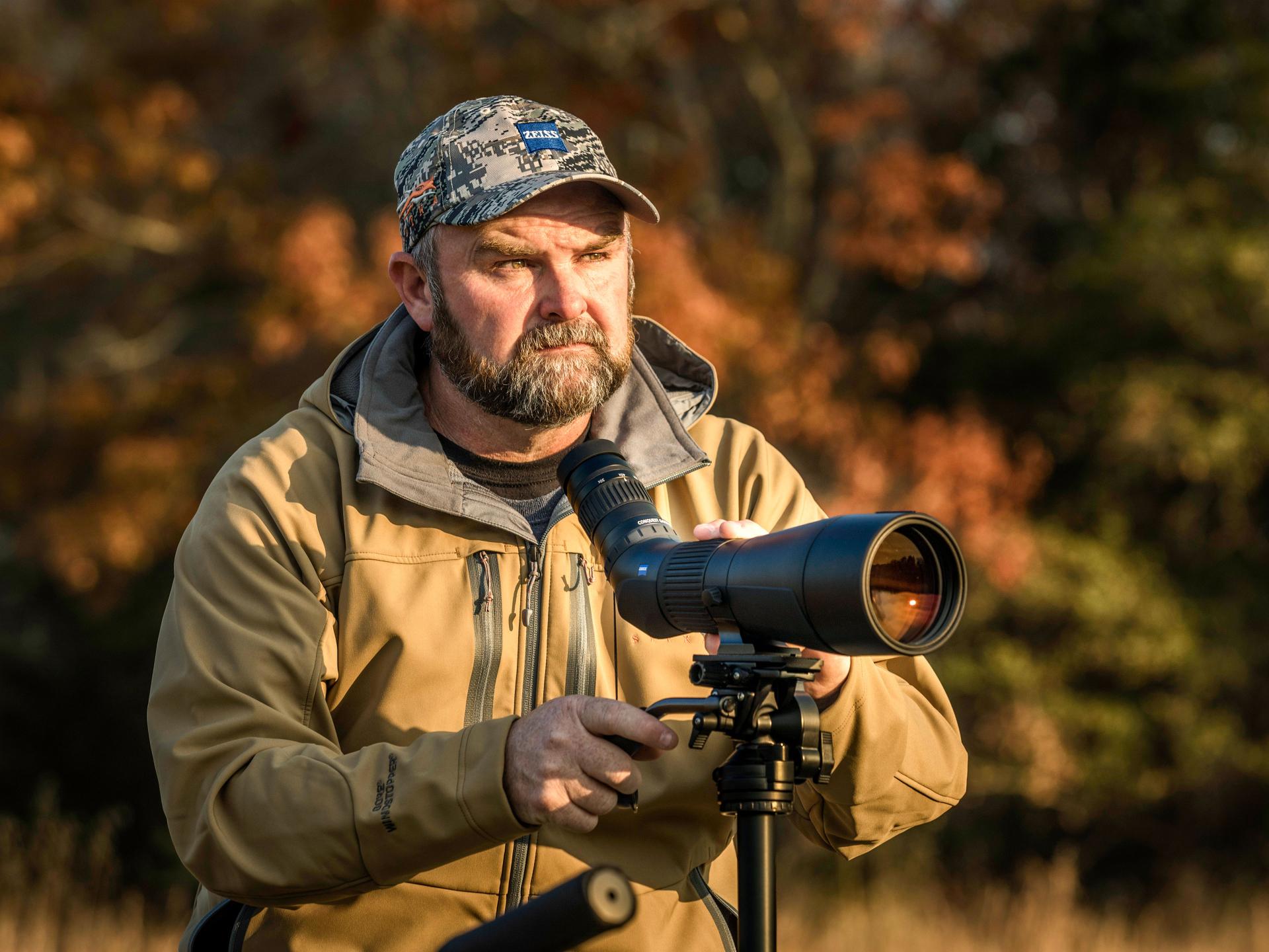 A man wearing outdoor gear adjusts a ZEISS spotting scope mounted on a tripod in a natural, autumn setting.