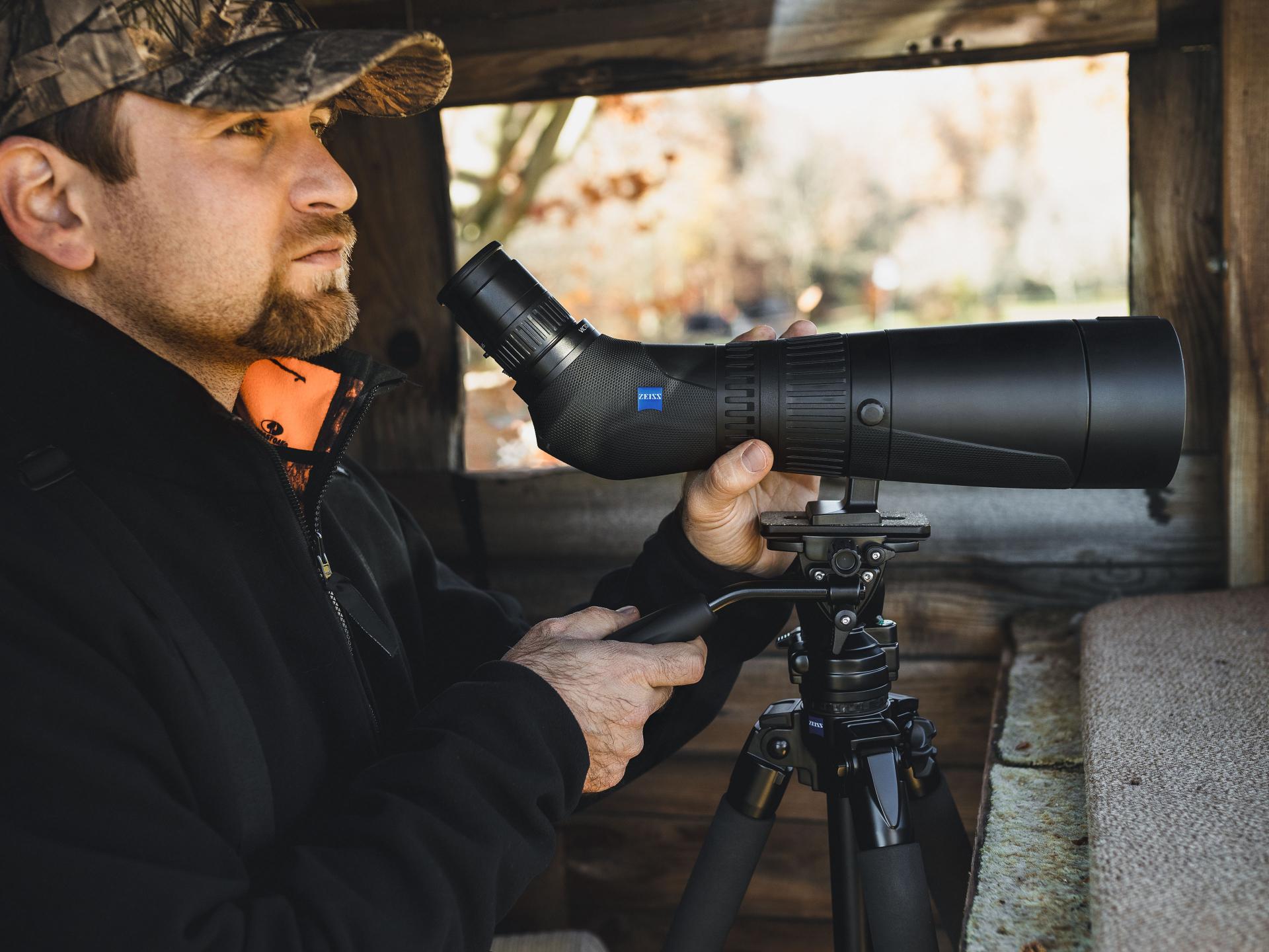 A man in outdoor gear uses a ZEISS spotting scope mounted on a tripod inside a wooden observation shelter.