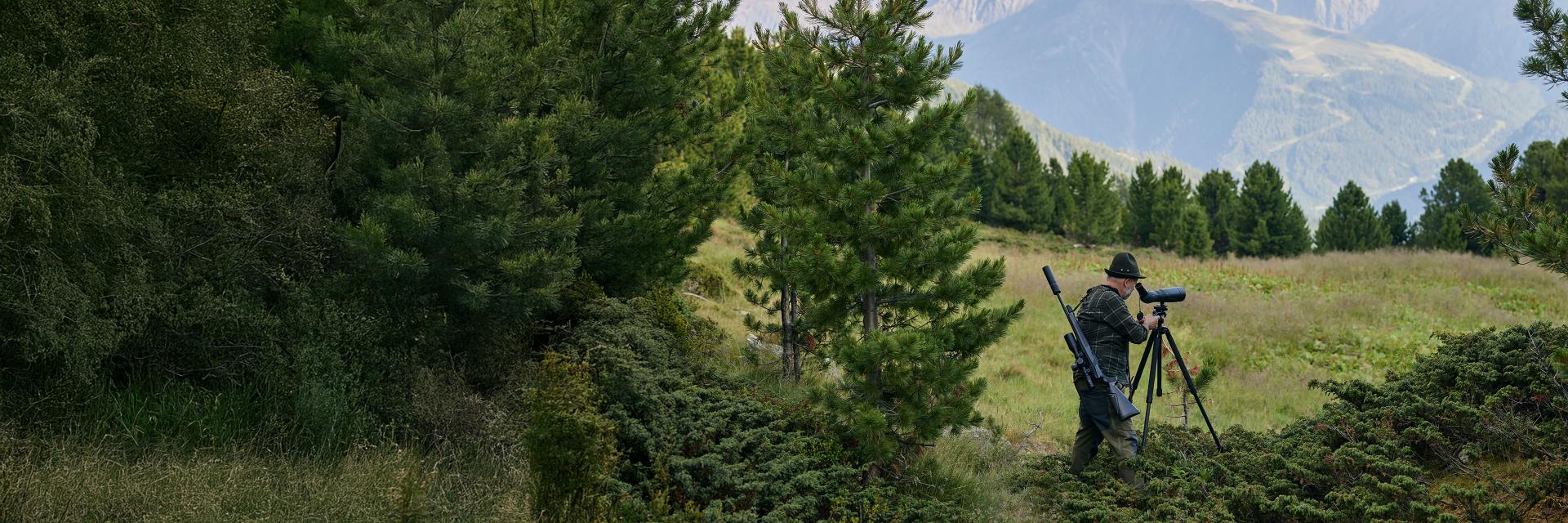Man using a ZEISS spotting scope on a tripod in a forested outdoor setting.