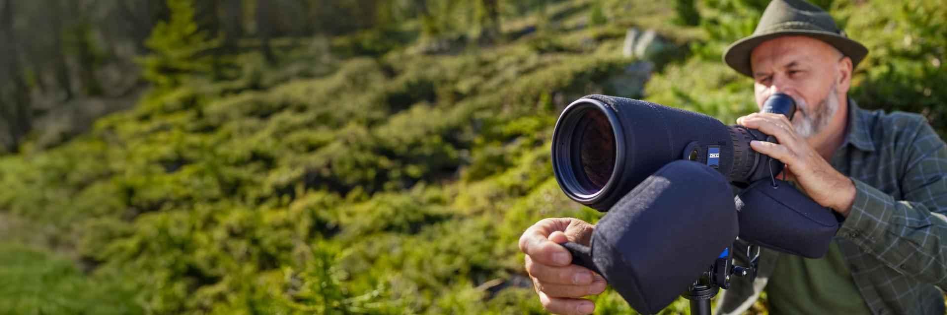 Person in camouflage clothing using a ZEISS spotting scope on a tripod in a grassy outdoor setting.