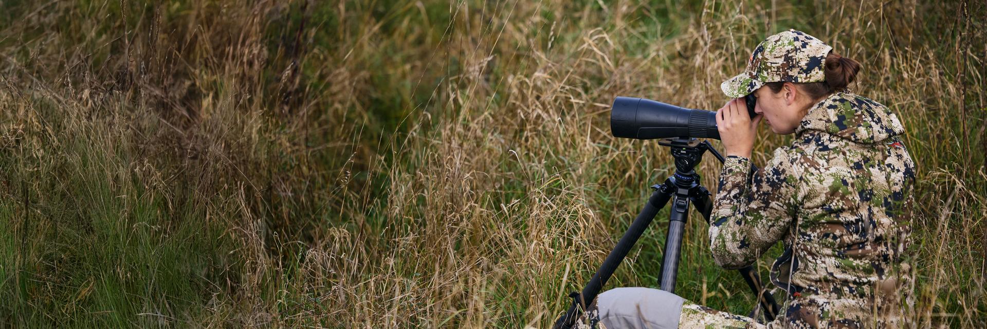 Person in camouflage clothing observes through a large ZEISS spotting scope mounted on a tripod in a grassy outdoor setting.