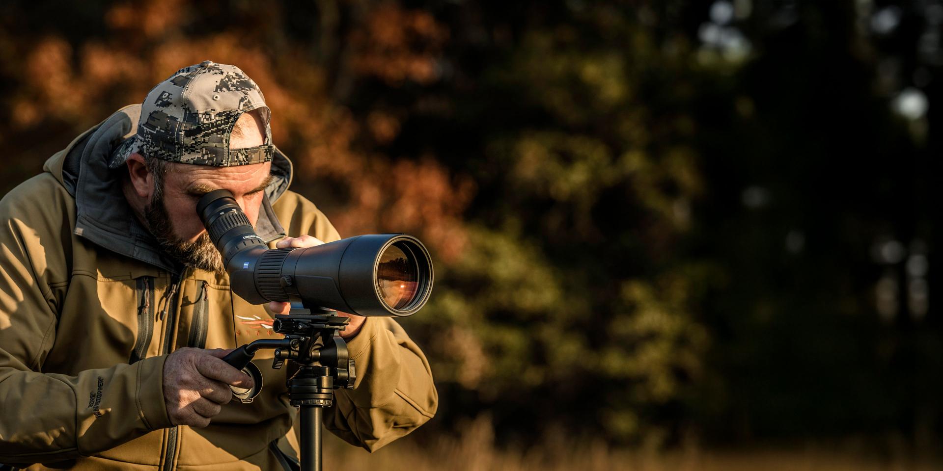 A man in outdoor gear is looking through a ZEISS spotting scope mounted on a tripod in a natural setting.
