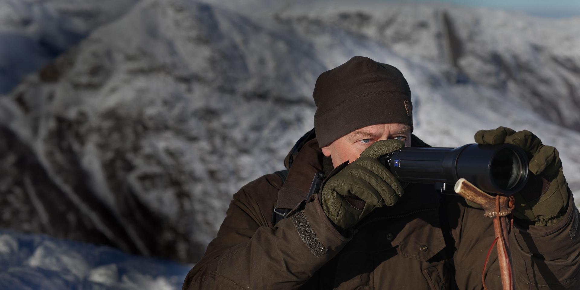 A man in winter clothing uses a pair of ZEISS binoculars while observing a snow-covered mountainous landscape.