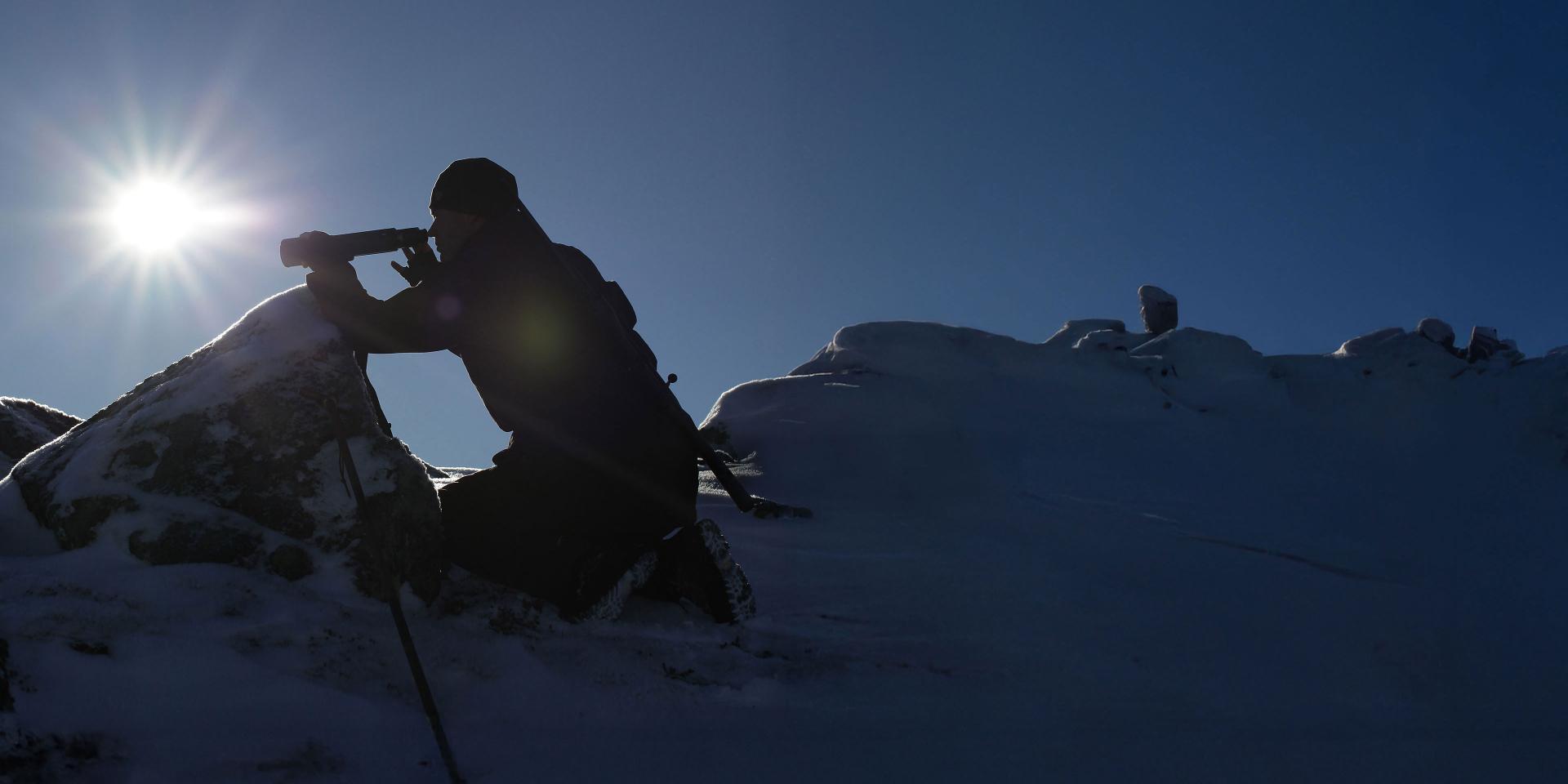 A person silhouetted against the sun uses a ZEISS spotting scope in a snowy mountain landscape.