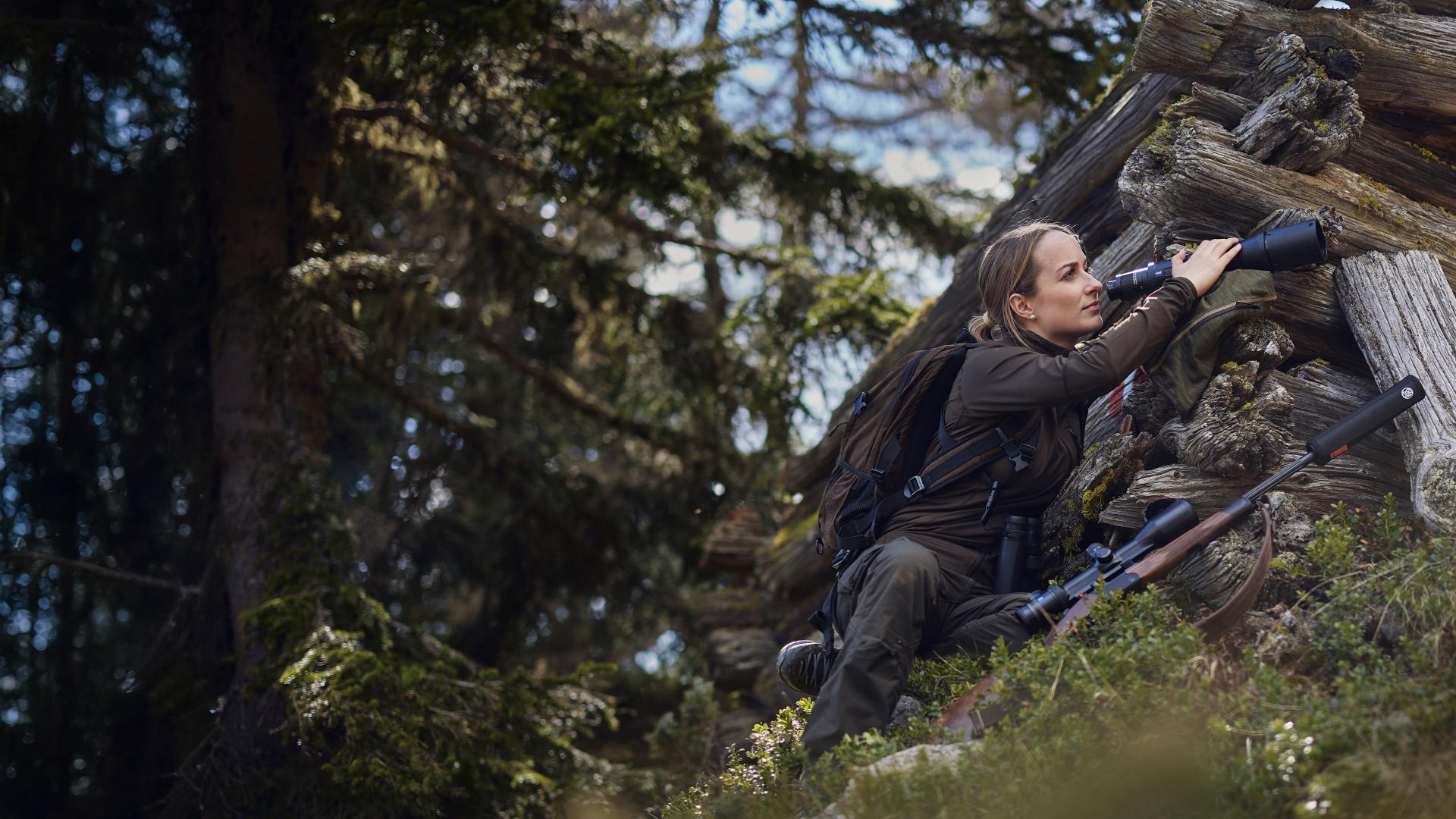 A woman in outdoor gear uses ZEISS binoculars while observing wildlife in a forested area next to a log pile.