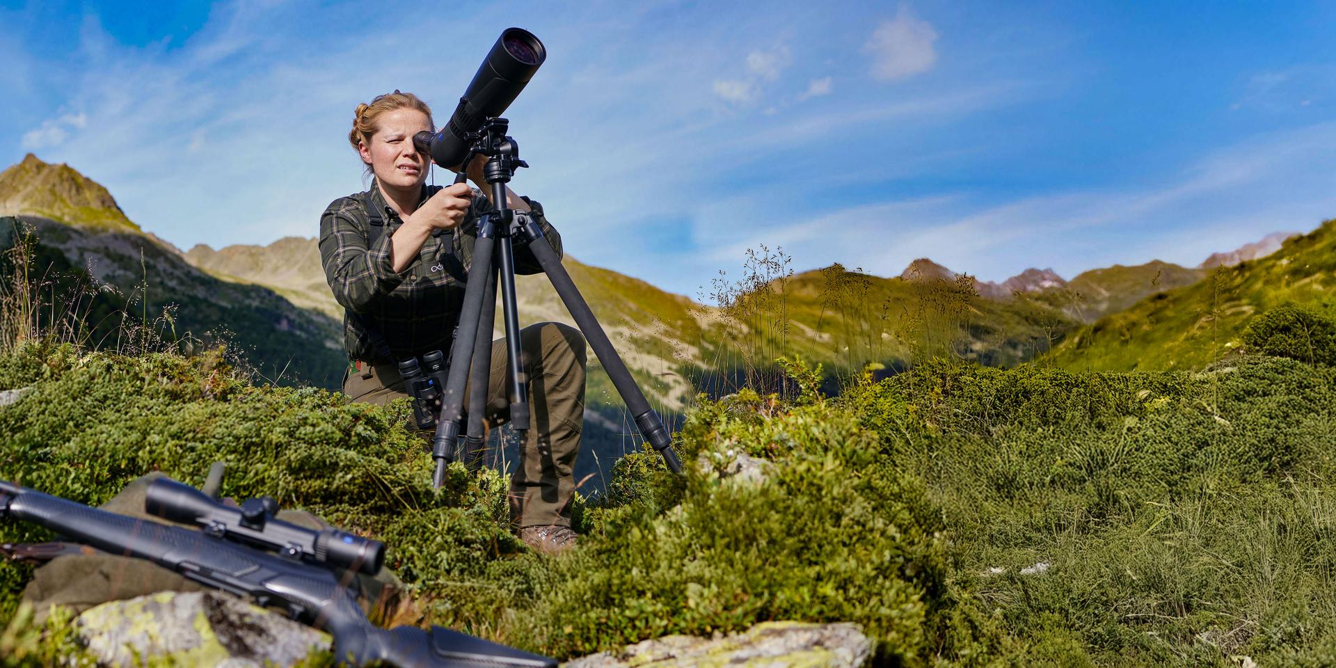 A person is adjusting a ZEISS spotting scope mounted on a tripod in a mountainous outdoor landscape.