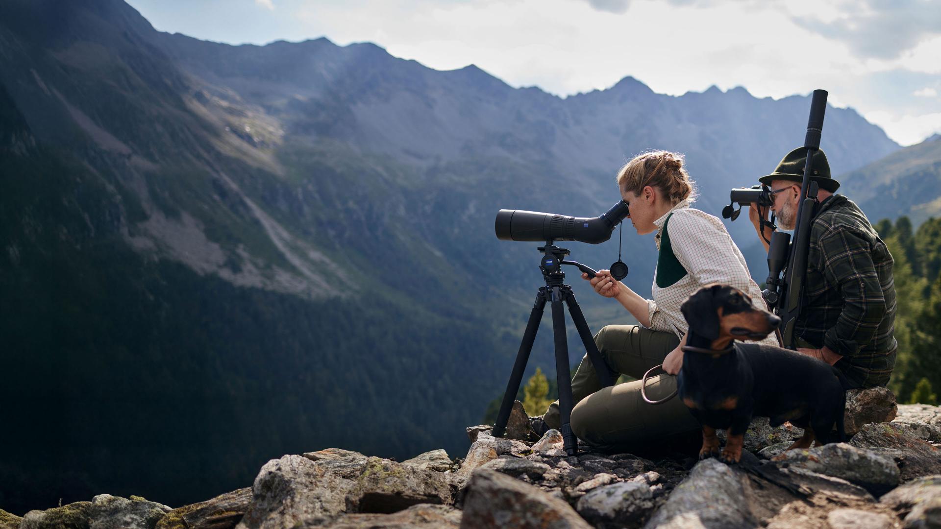 Two people on a mountain ledge use ZEISS spotting scopes and binoculars to observe the landscape, accompanied by a dog.