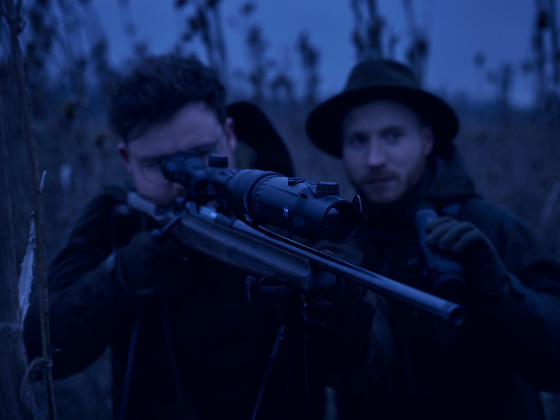 Two men are outdoors in low light, with one aiming a rifle equipped with a ZEISS riflescope while the other observes closely.