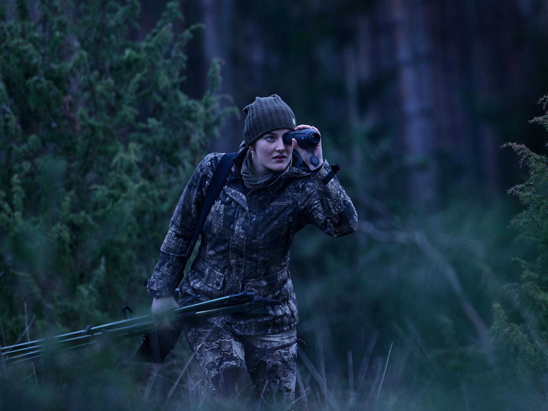 A person in full camouflage gear uses ZEISS optics while walking through a dense forest.