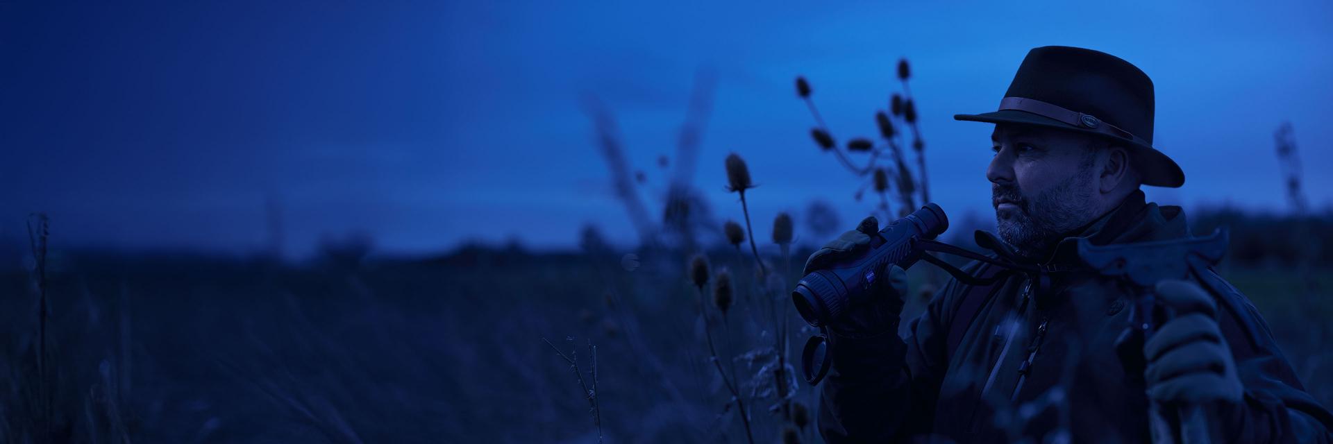 A man in outdoor gear and a hat observes the landscape through ZEISS binoculars in a field at dusk.