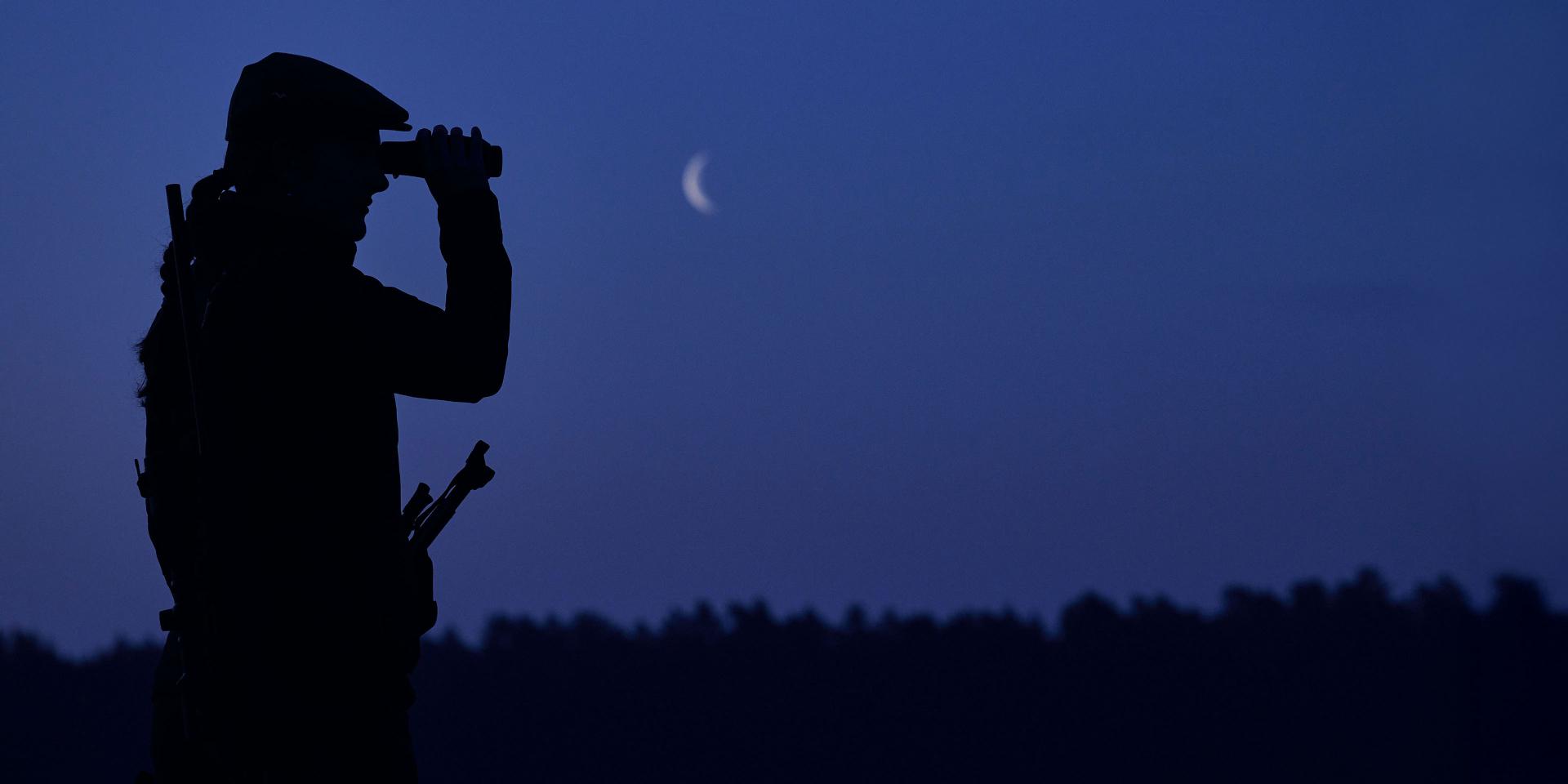 Silhouette of a person using ZEISS binoculars at dusk with a crescent moon in the sky and a forested landscape in the background.