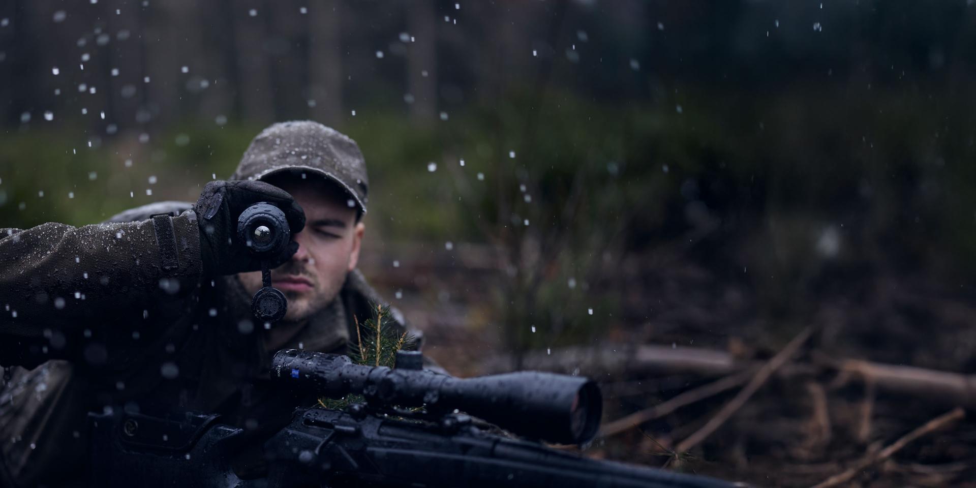A man in camouflage clothing aims through a ZEISS riflescope in a forest during rainfall.