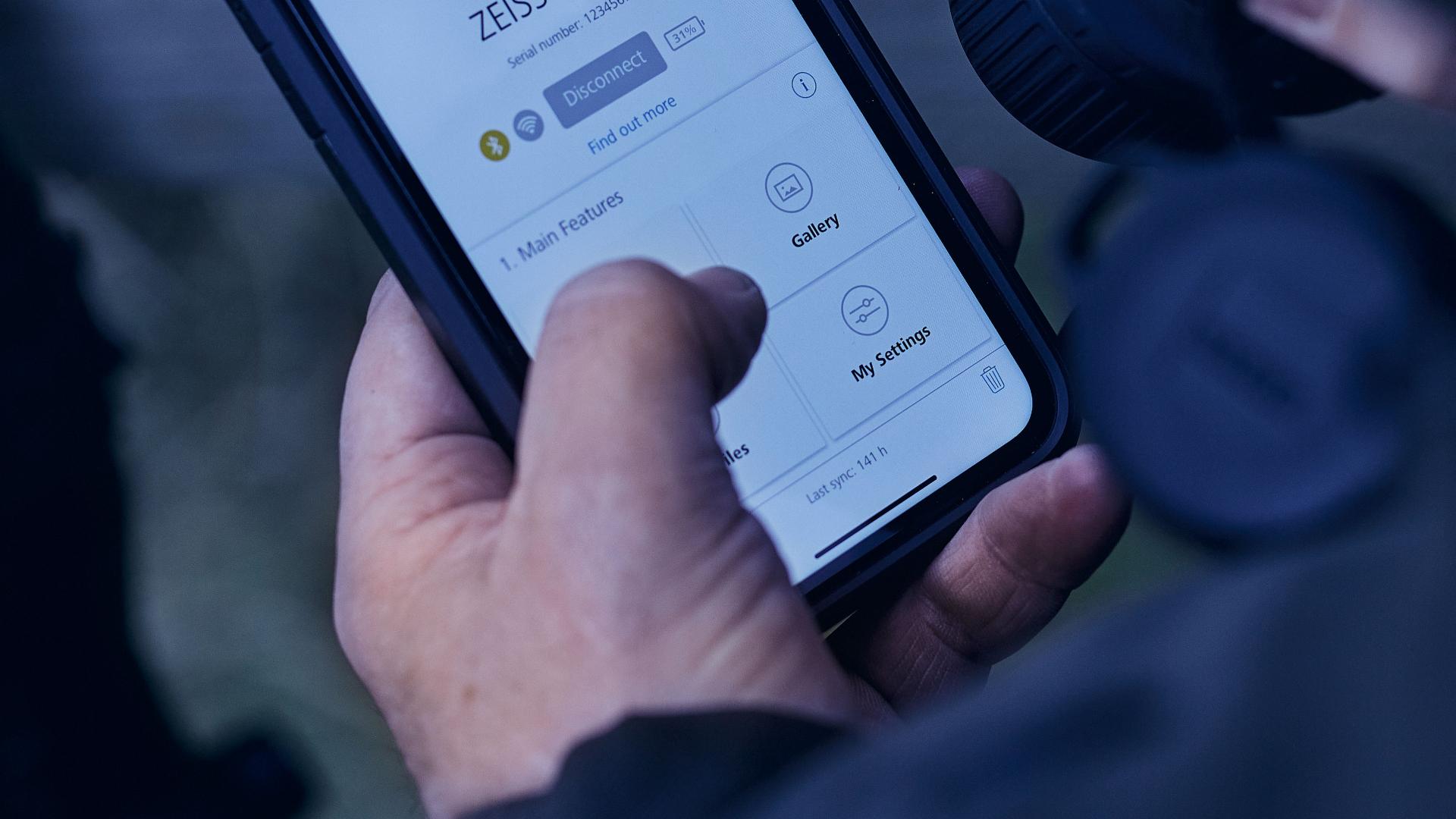 A person holds a smartphone displaying the ZEISS app interface with options like "Gallery" and "My Settings."