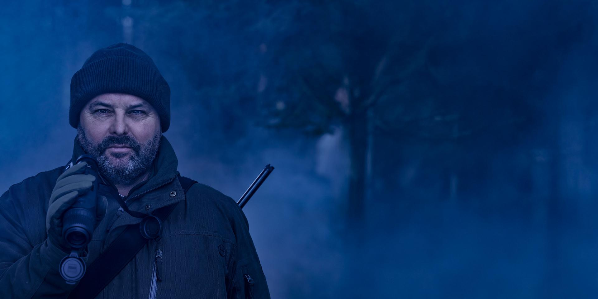 A man dressed in outdoor gear stands in a misty forest holding a rifle equipped with a ZEISS optical device.