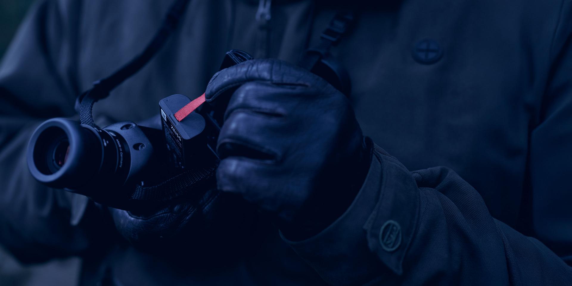 Gloved hands adjusting a camera equipped with a ZEISS lens in an outdoor setting.