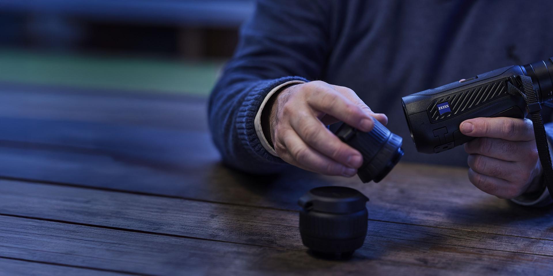 A person is assembling a ZEISS camera lens onto a ZEISS optical device on a wooden table.