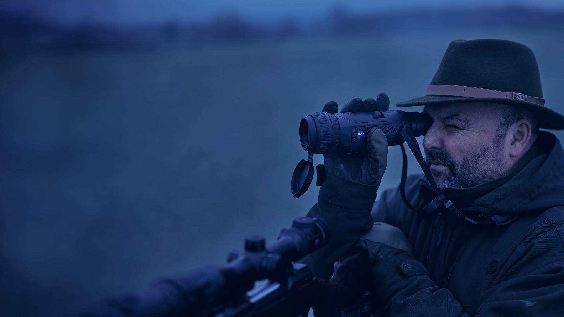 A man wearing outdoor gear and a hat uses a ZEISS thermal imaging device while aiming with a rifle in a low-light environment.