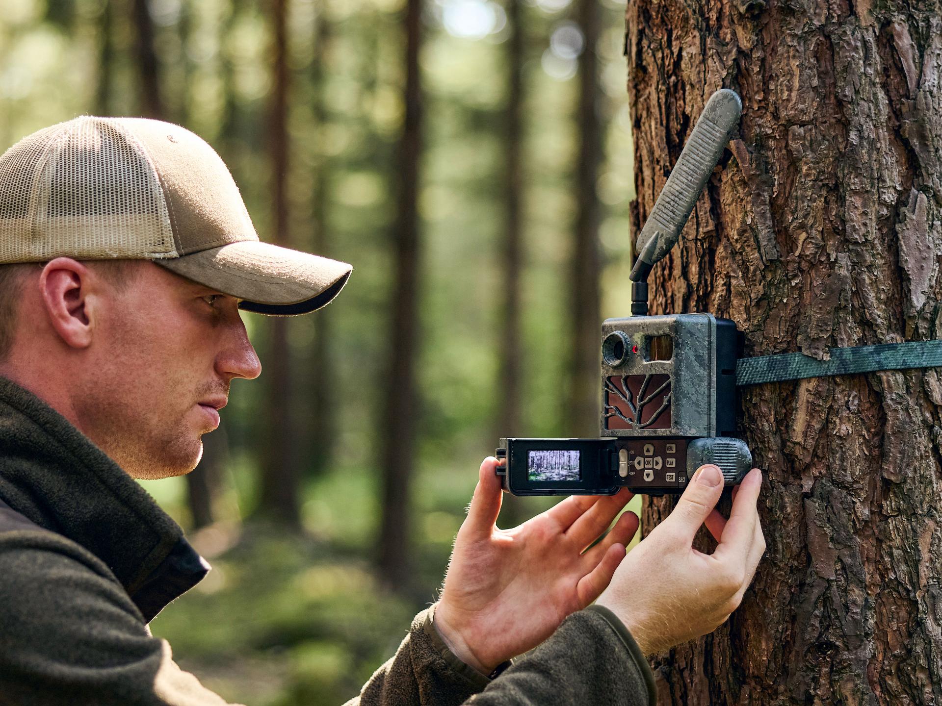 A man in outdoor clothing sets up a ZEISS trail camera with an external antenna on a tree in a forest.