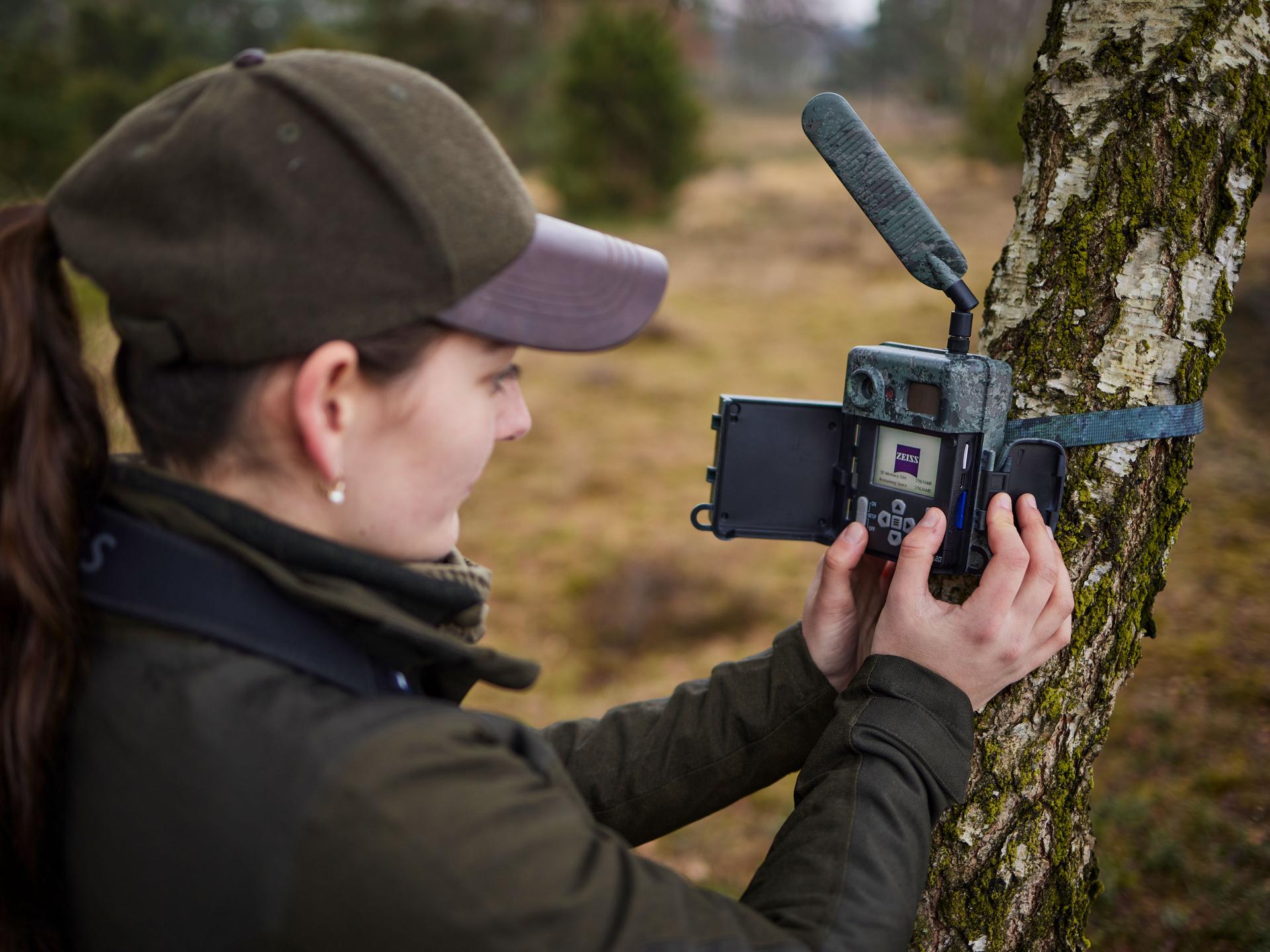 A person mounts and configures a trail camera on a tree in a forested area using ZEISS equipment.