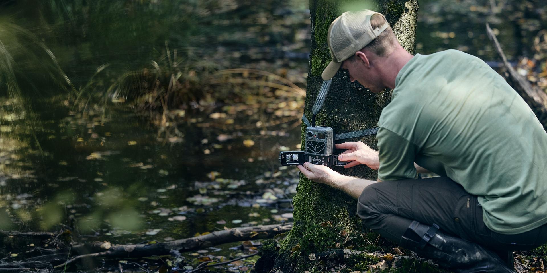 A man in outdoor gear sets up a ZEISS trail camera on a tree beside a forest pond.