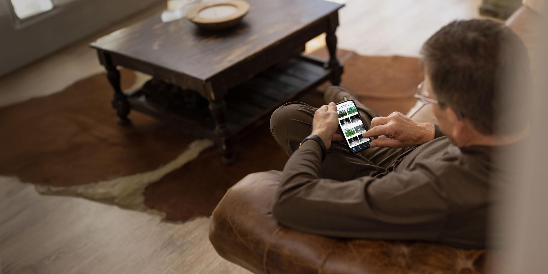 A man is sitting on a leather couch using a smartphone to view wildlife images, with a wooden coffee table and cowhide rug in the background.