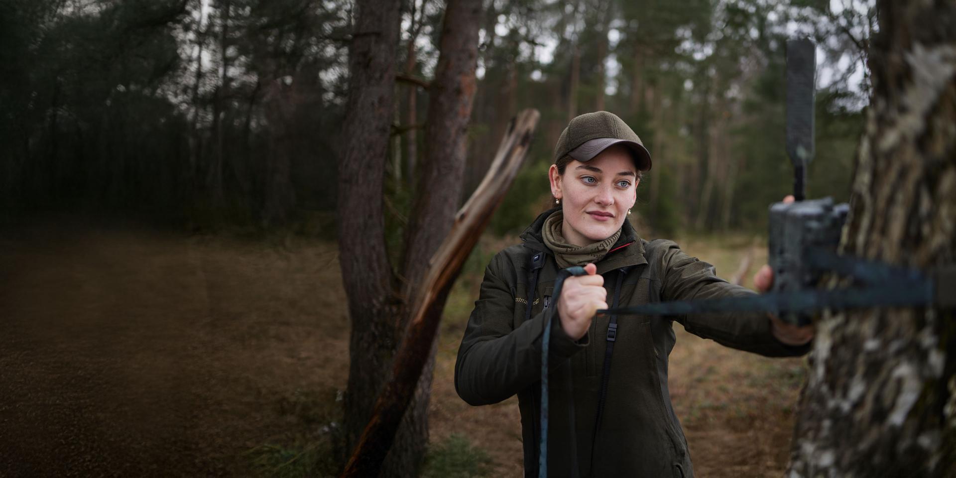 A woman in outdoor clothing is mounting a trail camera to a tree in a forested area, using ZEISS equipment.