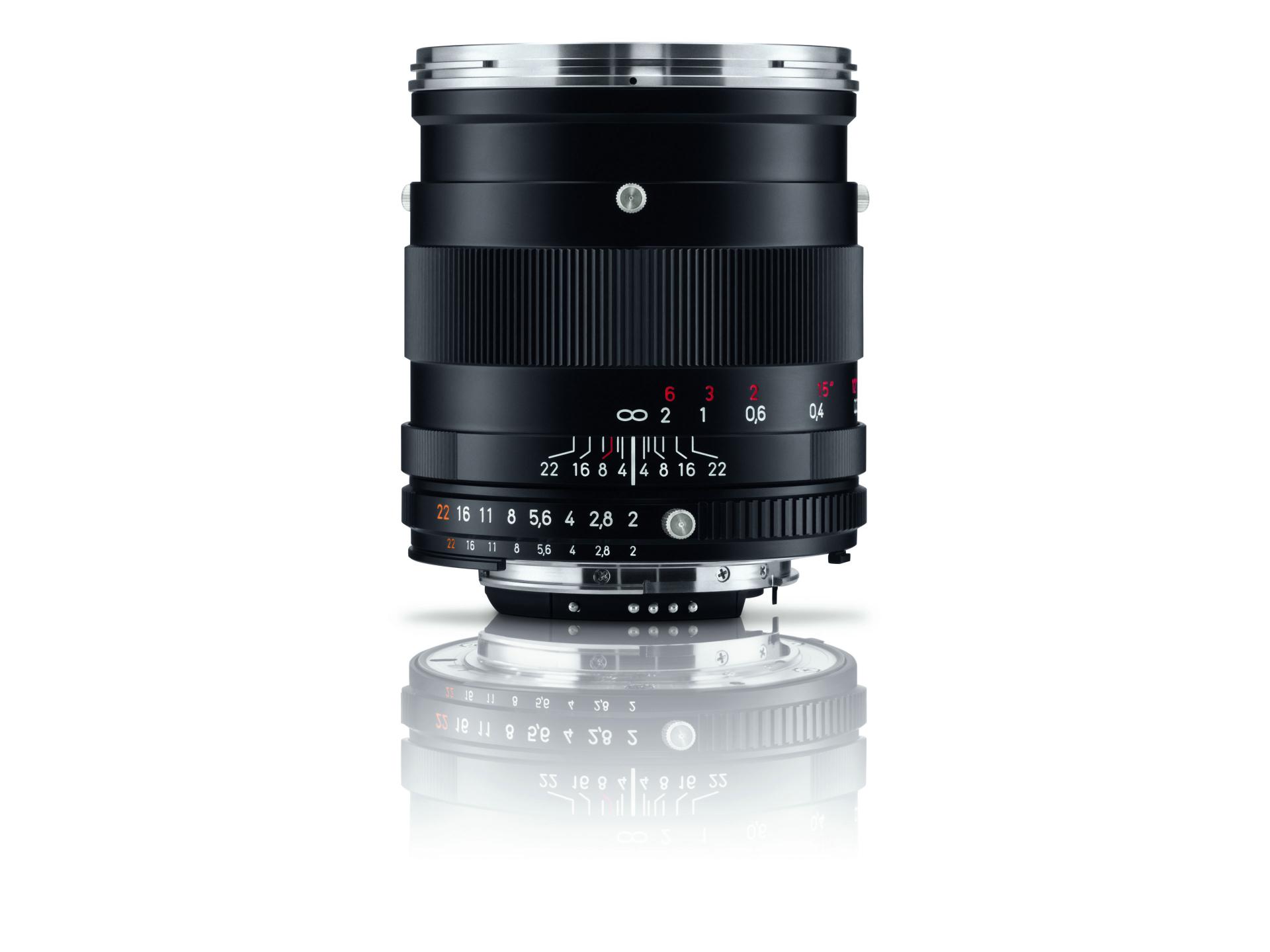 Black ZEISS camera lens with manual focus and aperture rings, displayed upright on a reflective white surface.
