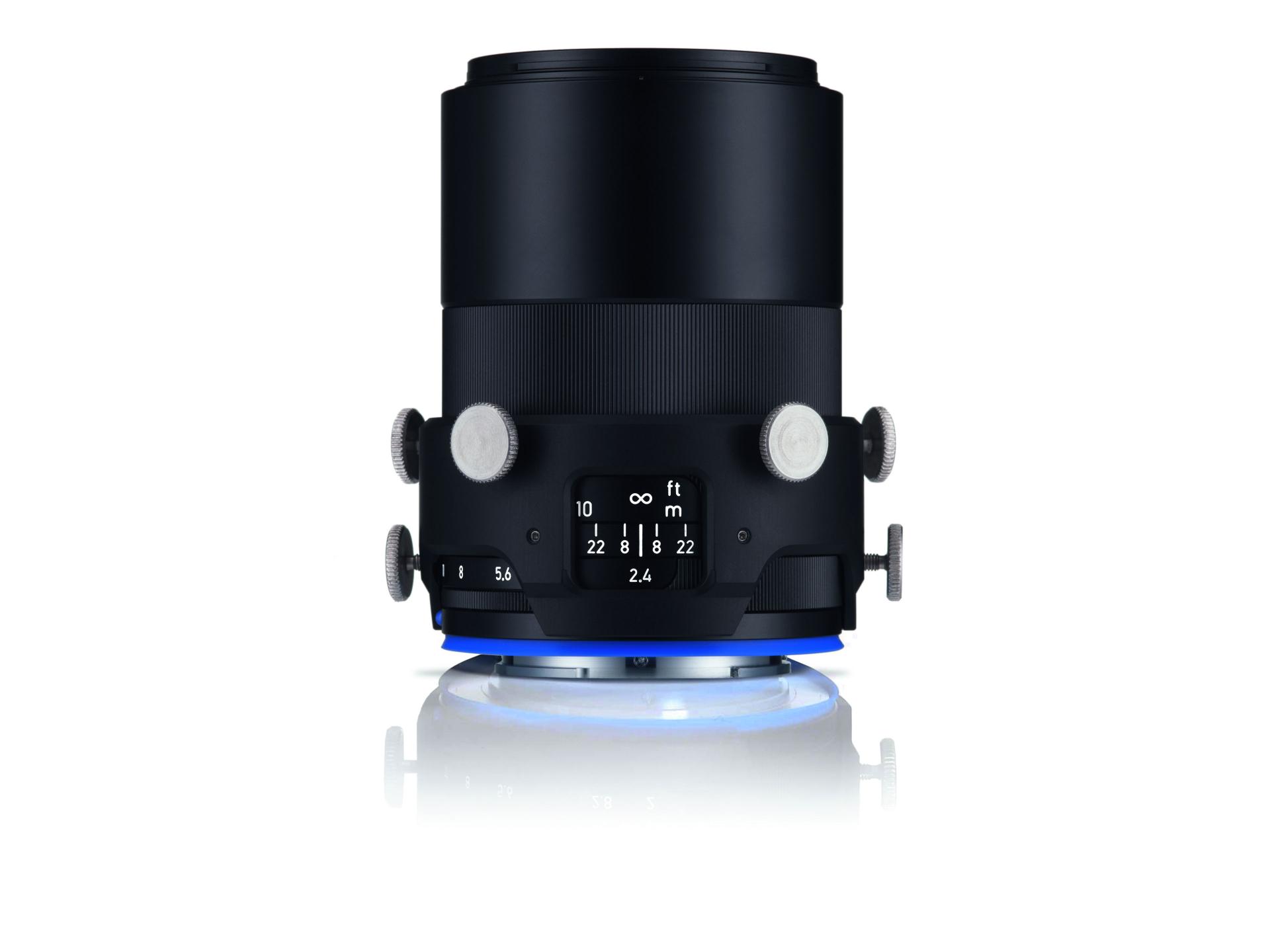 Black ZEISS camera lens with manual control knobs and distance markings, displayed upright on a reflective white surface.