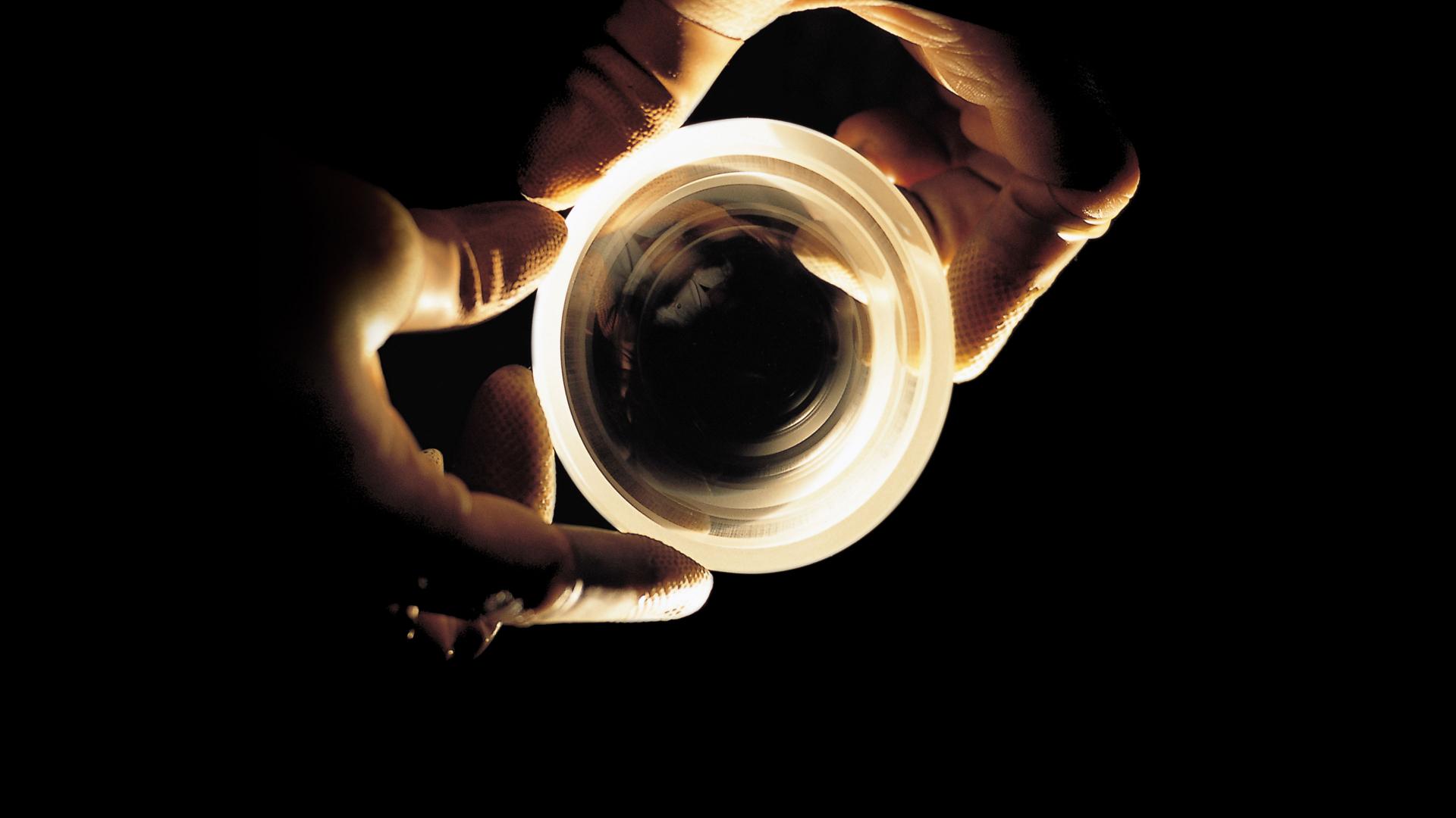 Two gloved hands holding a precision optical lens against a dark background.