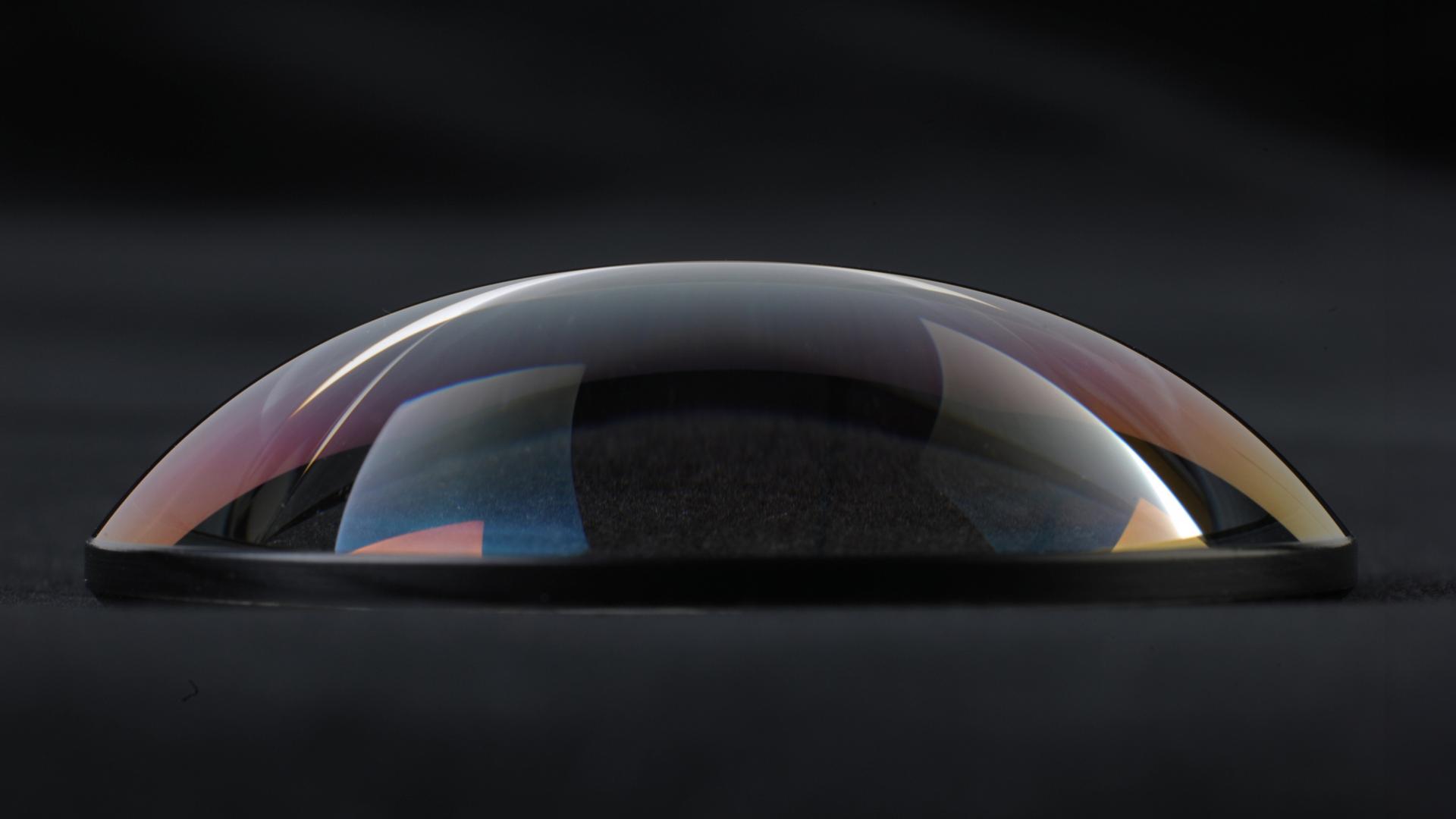 A close-up side view of a precision optical lens with a curved, reflective surface placed on a dark background.