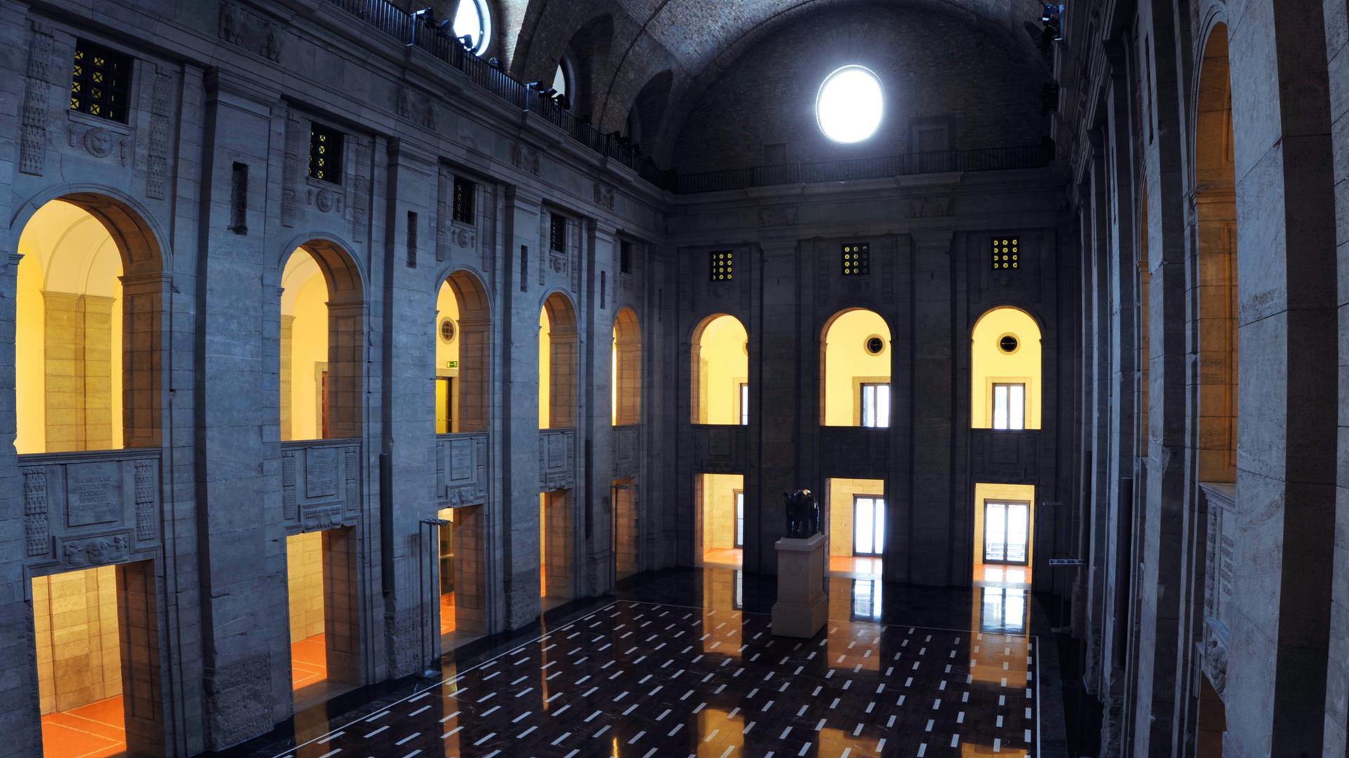 Interior view of a grand hall with tall arched windows, stone walls, and a polished dark floor with light reflections.