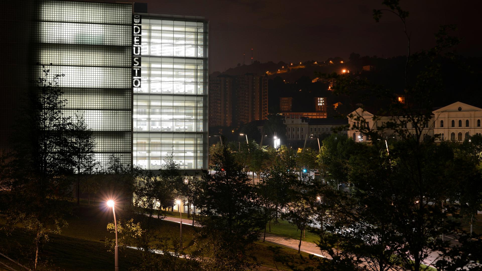 Modern glass building illuminated at night with the word "DEUSTO" displayed vertically on the facade, surrounded by trees and city lights in the background.