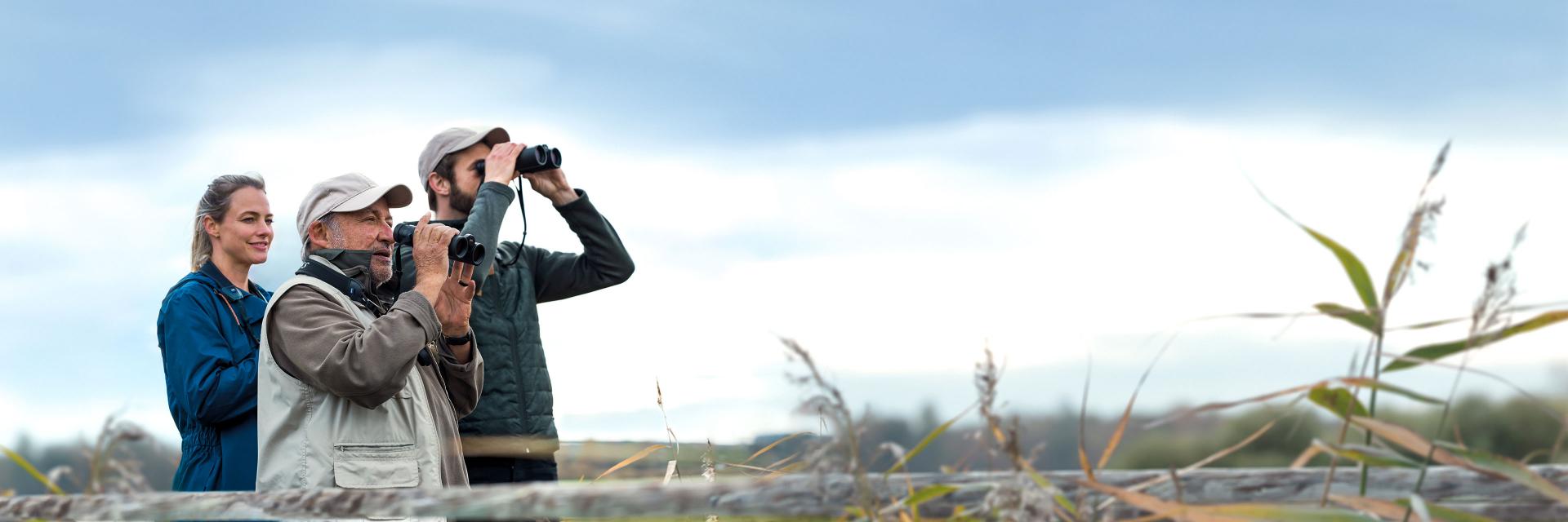 Three people outdoors using ZEISS binoculars to observe nature in a grassy, open landscape.