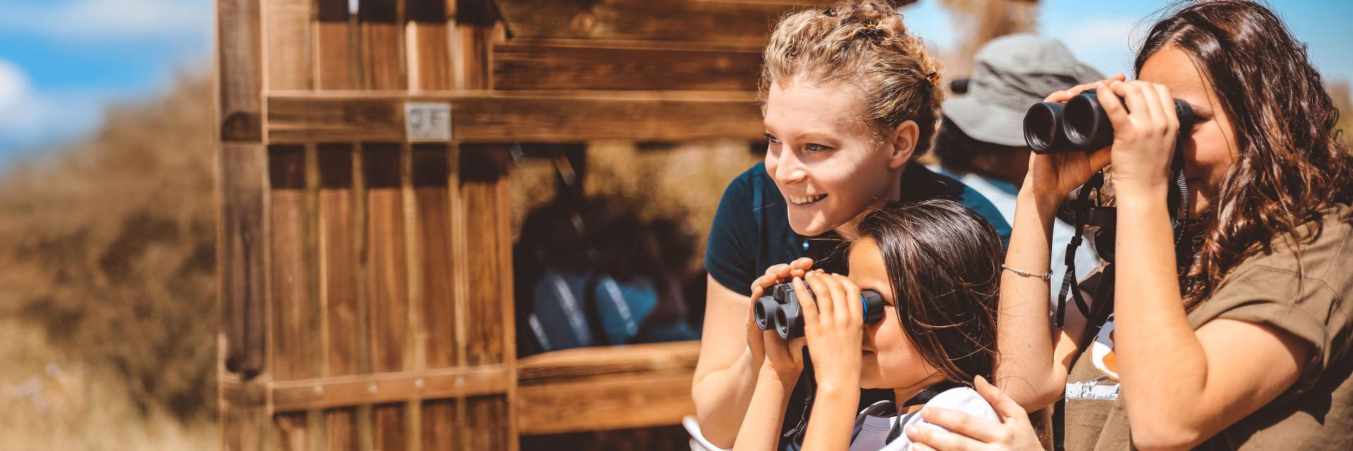 Three people, including a child, are outdoors using ZEISS binoculars to observe wildlife near a wooden lookout structure.