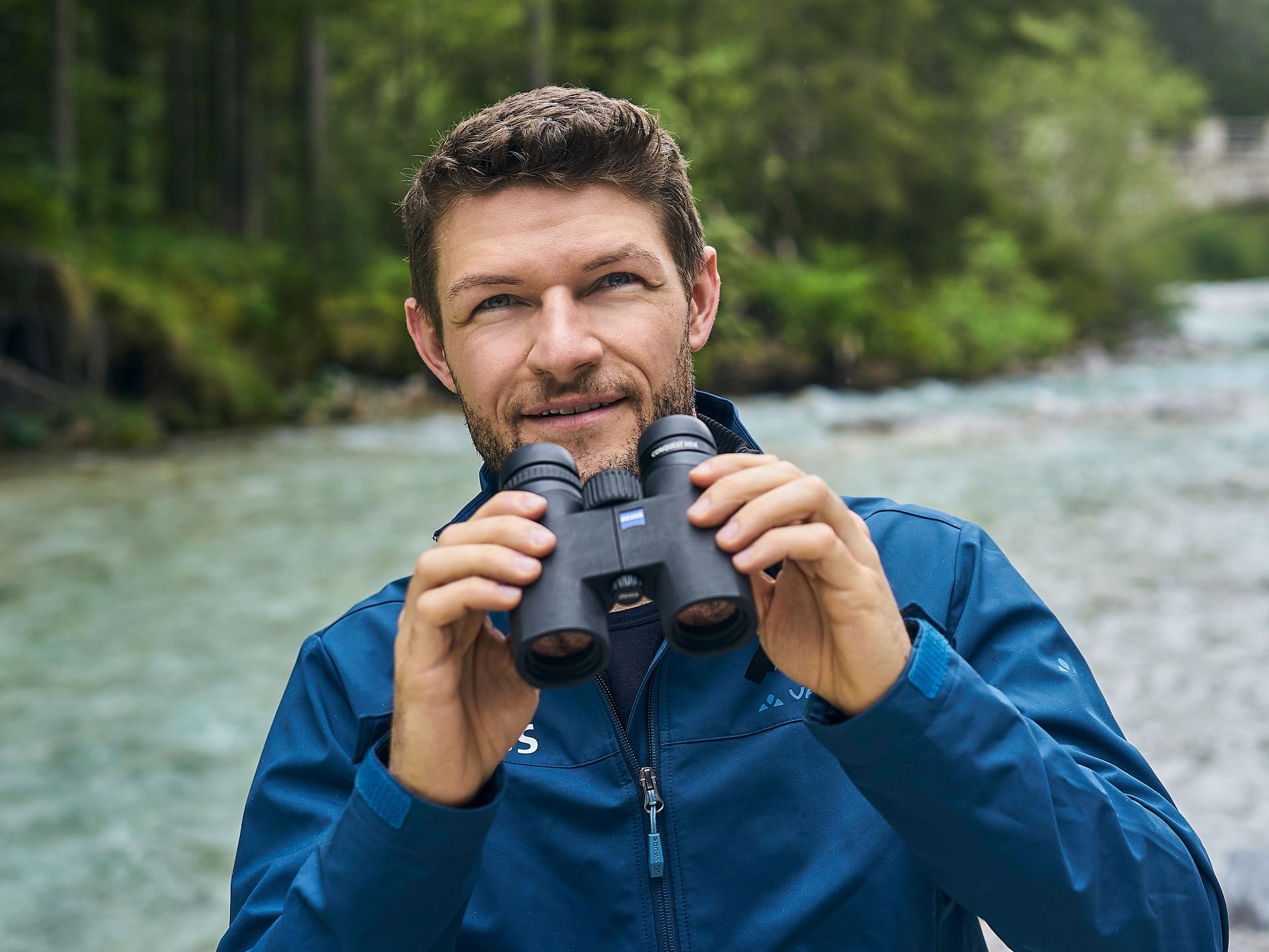 A man in a blue jacket holds a pair of ZEISS binoculars while standing beside a flowing river in a forested area.